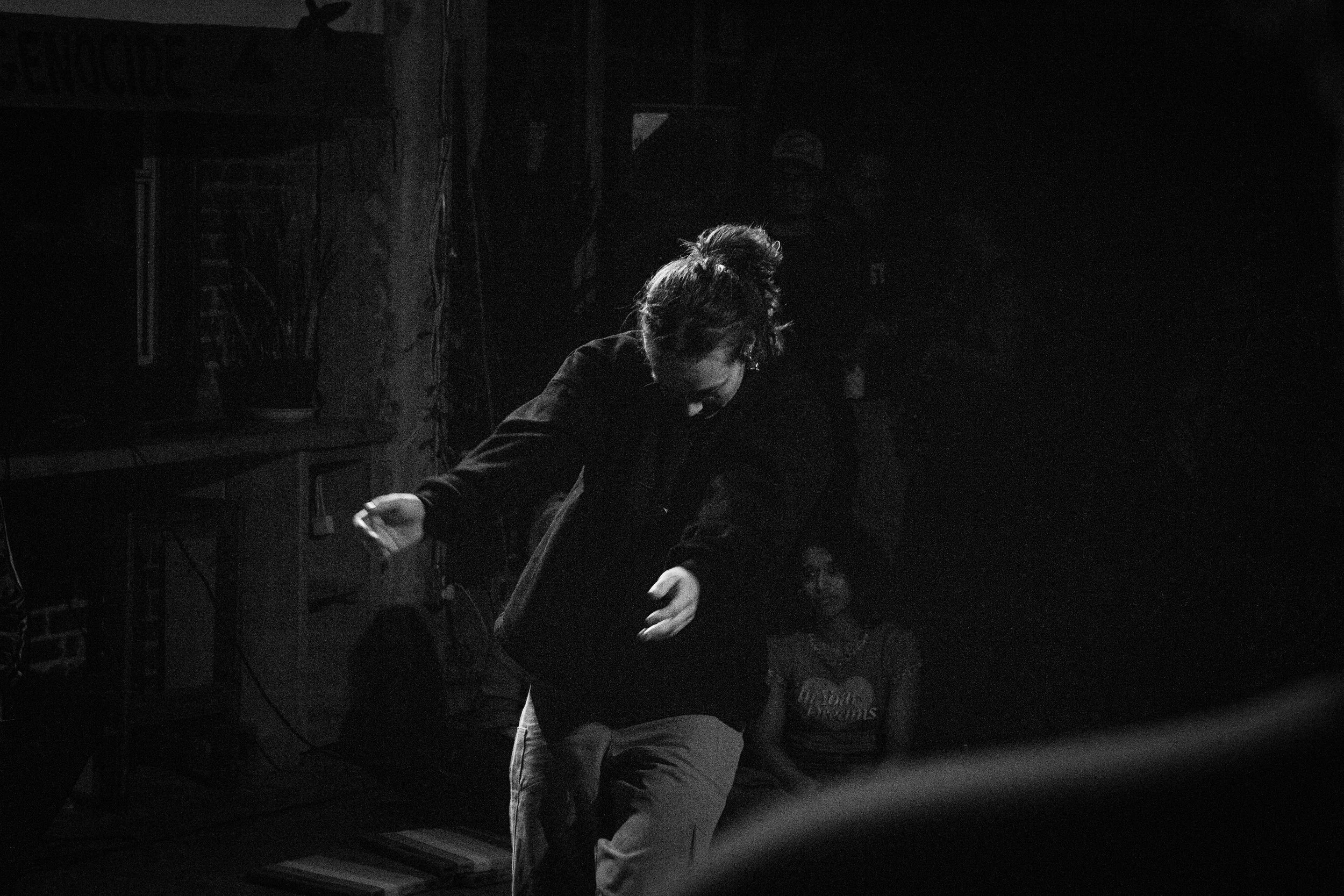 A person with curly hair in a bun, wearing glasses and dark clothing, leaning over and smiling, with a person in a striped shirt sitting in front of them in a dimly lit room.