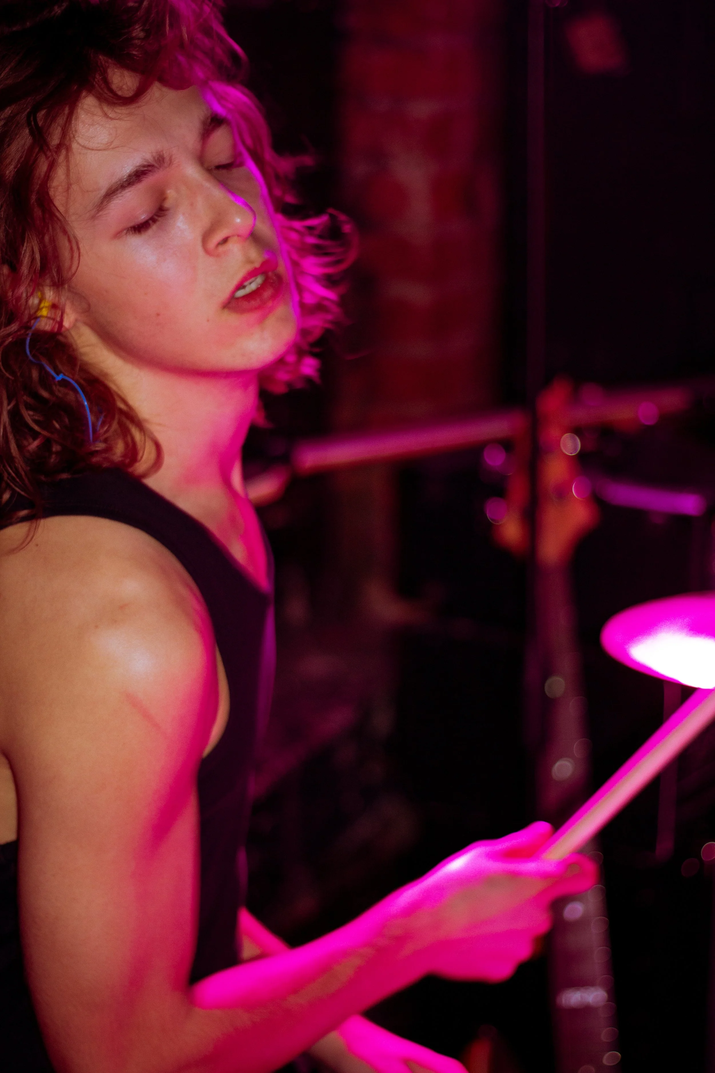 A young woman with curly hair and light skin is seen in a close-up shot, with her eyes closed and a focused expression, holding a glow stick or light tube in her hands. The scene is illuminated with pink lighting in a possibly dark environment, with 