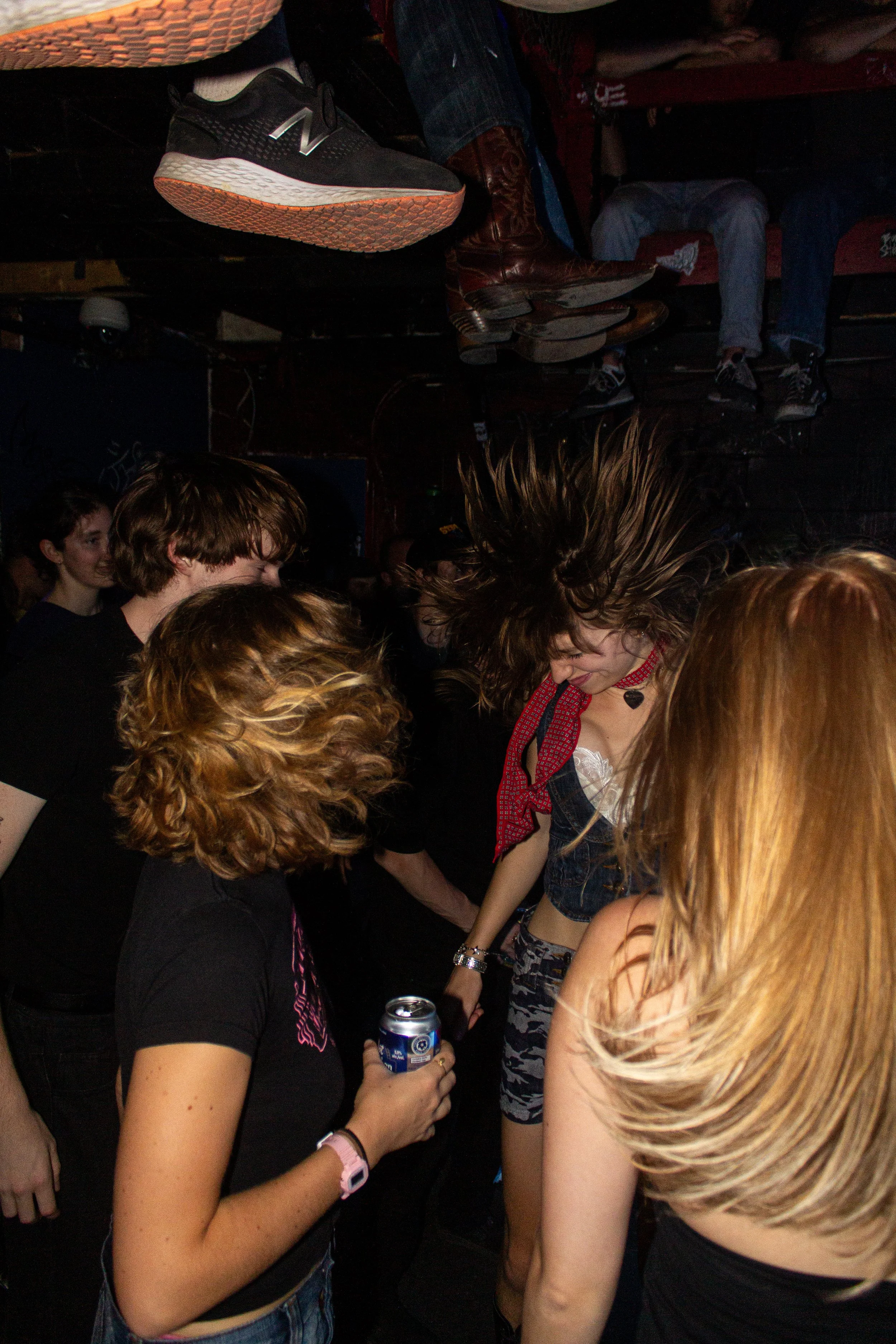 People at a party having a conversation, with one person holding a can of drink. The scene shows a crowd in a dimly lit space, with visible shoes and heads, and some women with long hair.