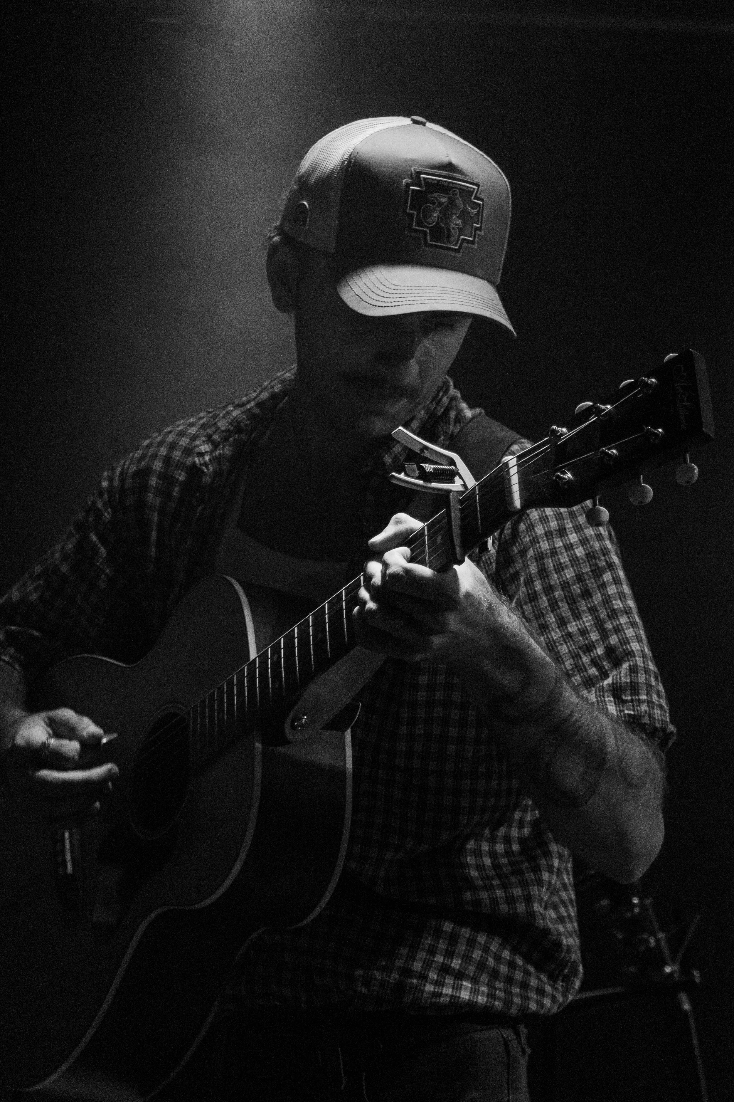 A man playing an acoustic guitar. He is wearing a baseball cap with a logo, a plaid shirt, and appears to be in a dimly lit room.