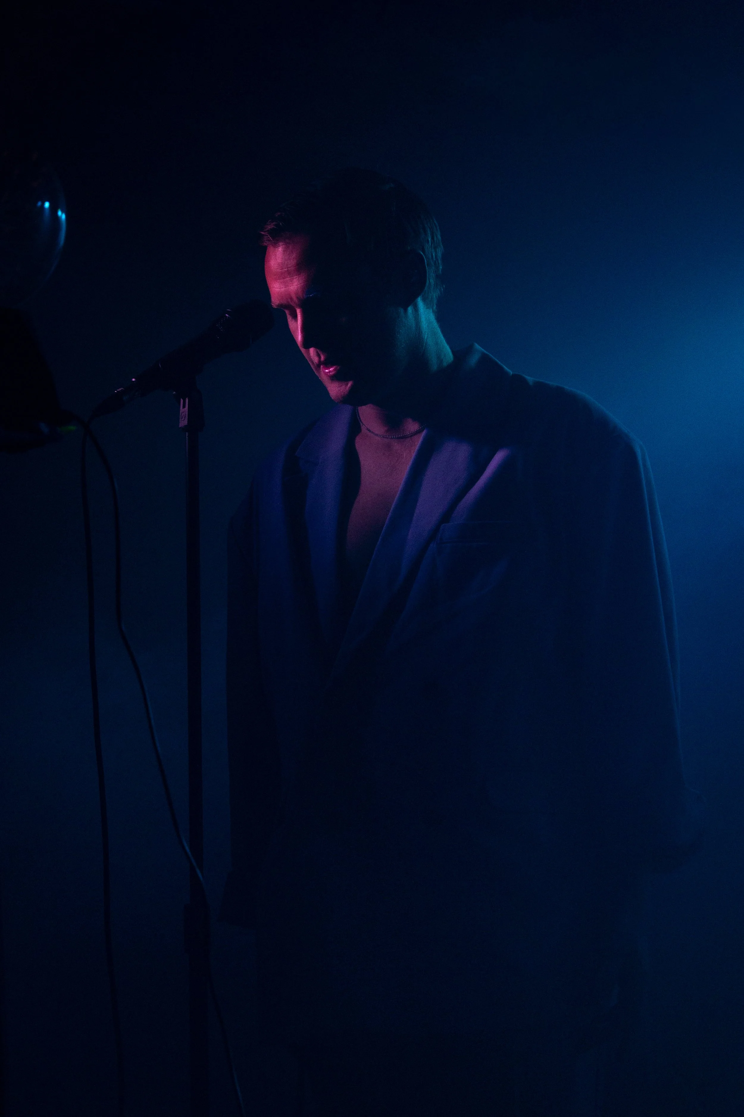 A male singer is performing with a microphone in front of him, illuminated by purple and blue stage lighting, with his eyes closed and a focused expression.