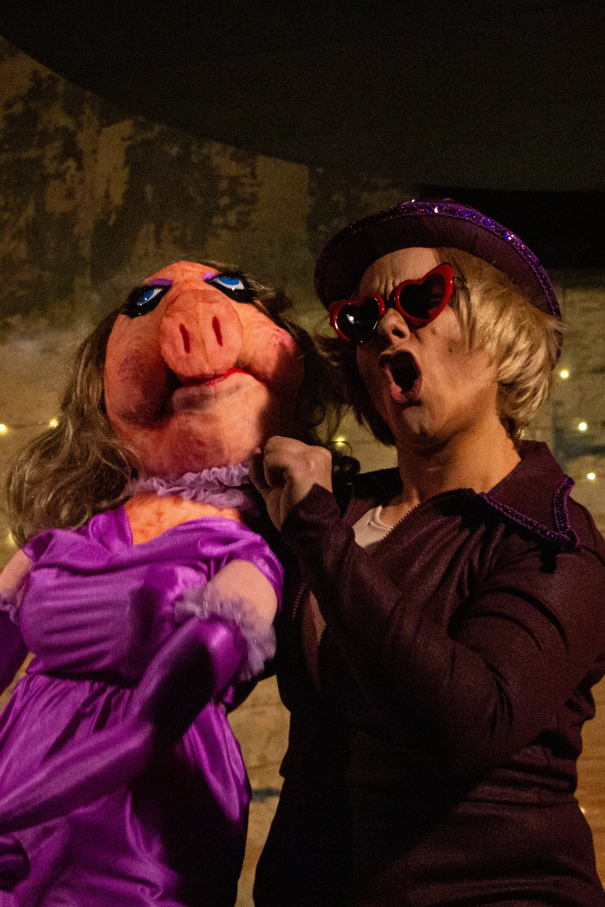 A person wearing heart-shaped sunglasses and a purple hat with purple glitter, next to a person in a pink pig costume with exaggerated facial features, in a festive setting with string lights.