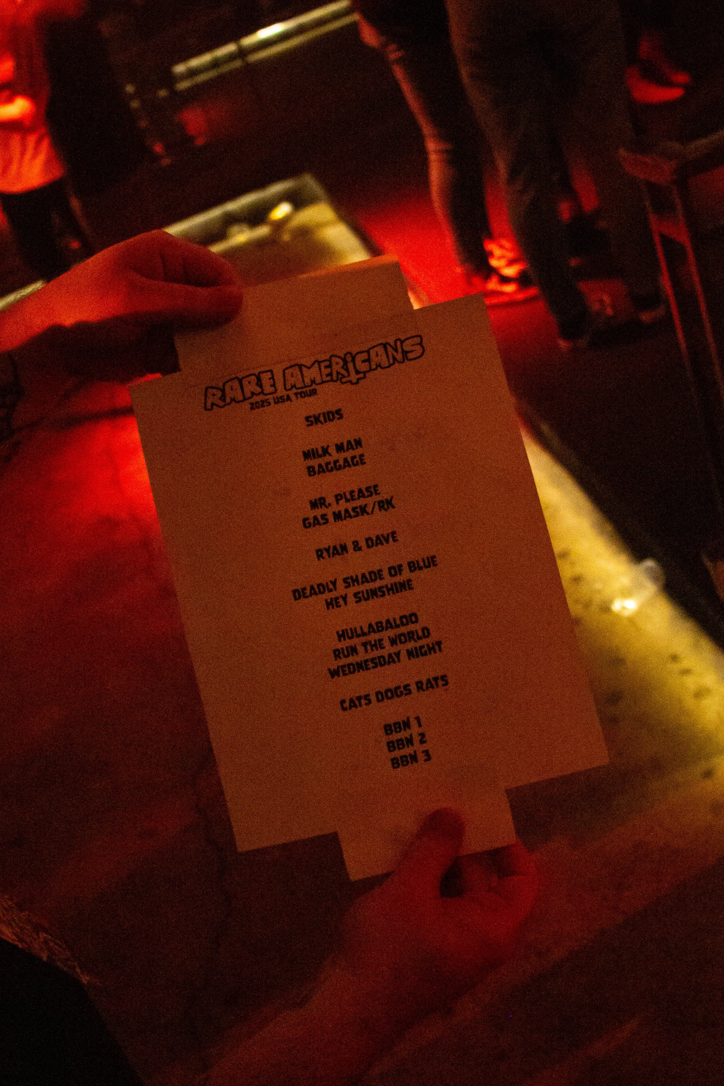 A person holding a song setlist for the Amo Americans 2023 USA tour, featuring titles such as "Skids," "Milk Man," "Baggage," and "Deadly Shade of Blue." The background shows a dimly lit indoor concert venue or bar with other people and musical equip