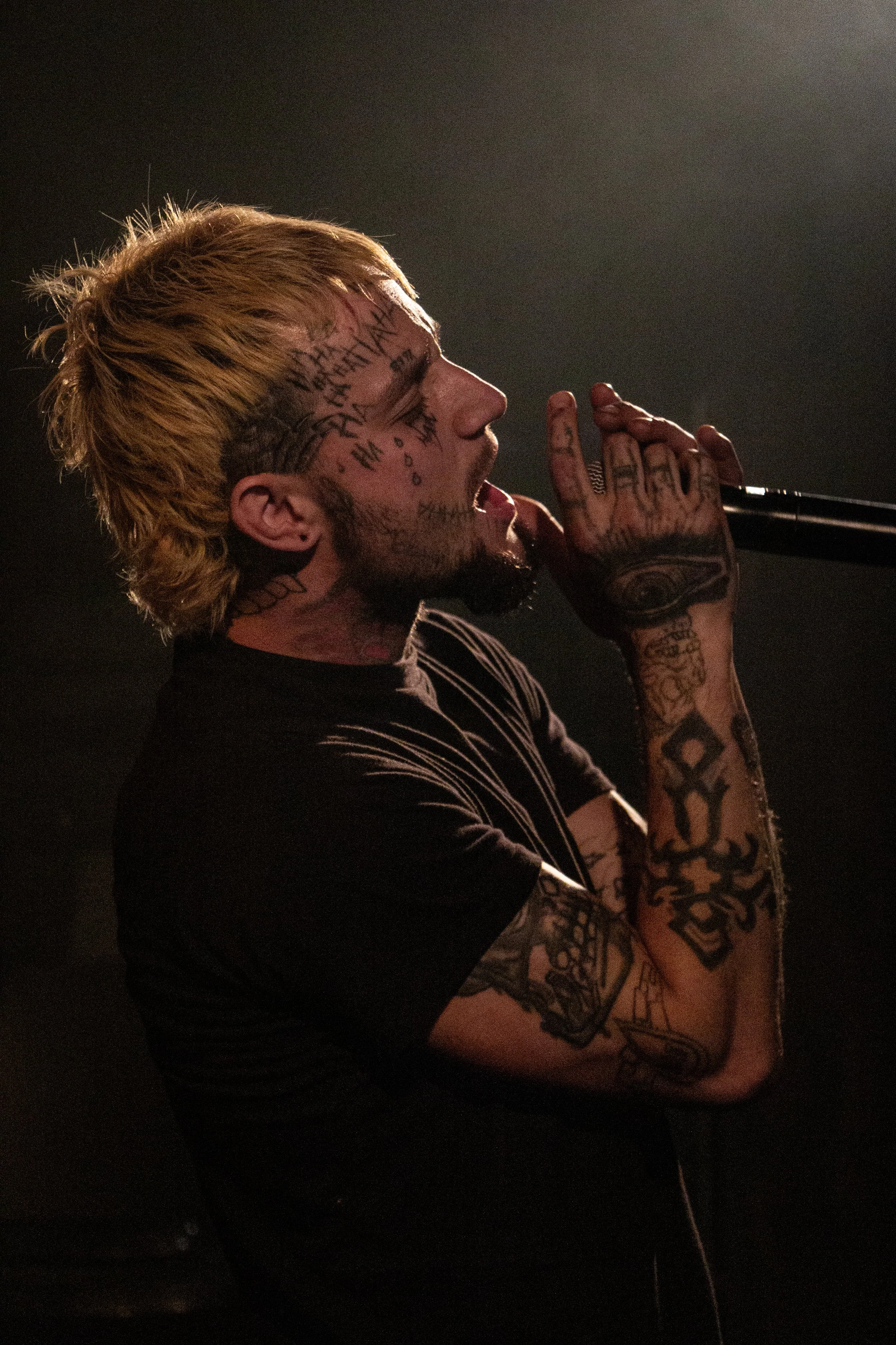 A young man with blonde hair and tattoos on his face and arms is singing into a microphone with his eyes closed.