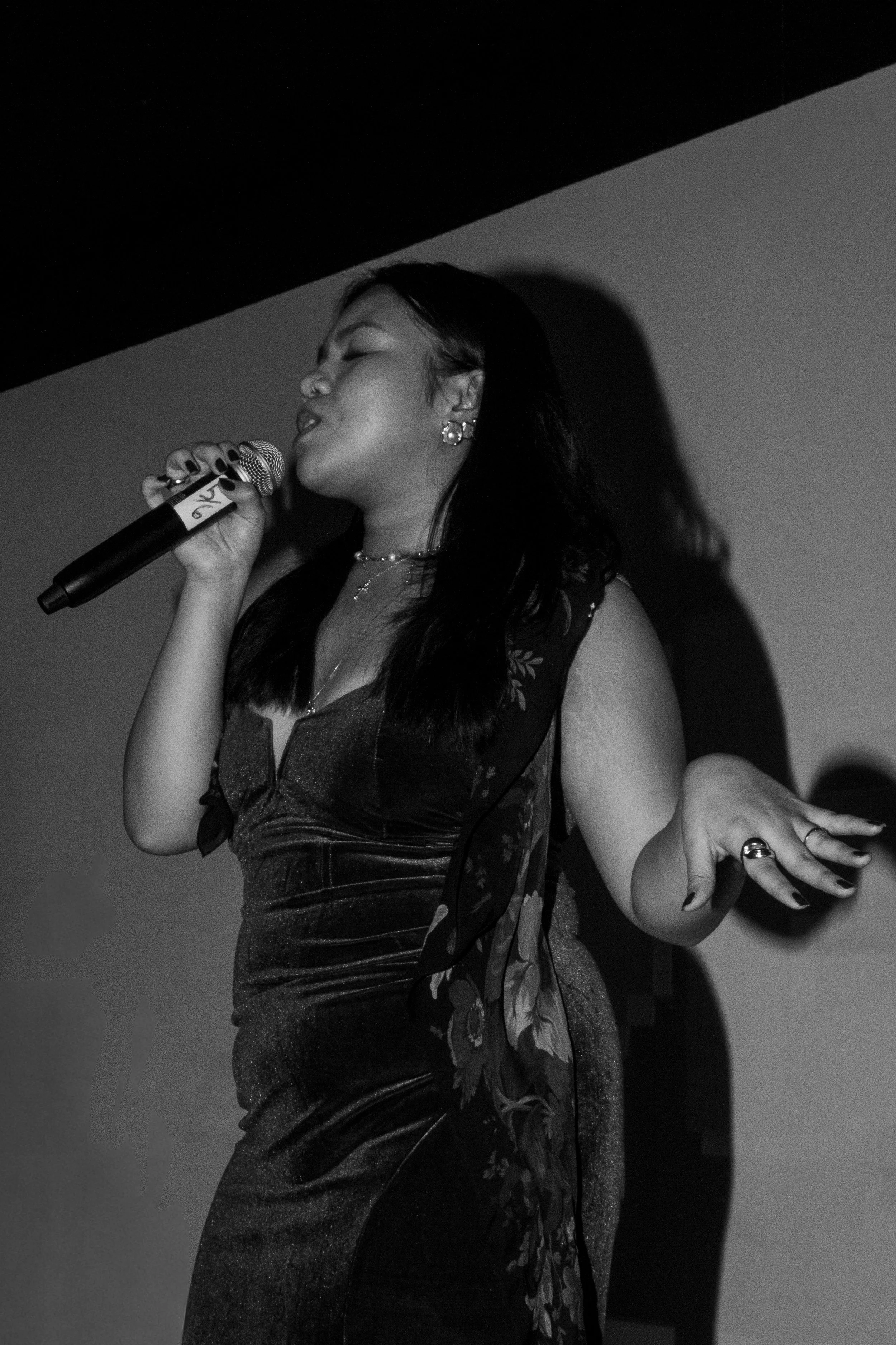 A woman singing into a microphone with her eyes closed, wearing earrings and a dress with floral patterns on the side, in black and white.