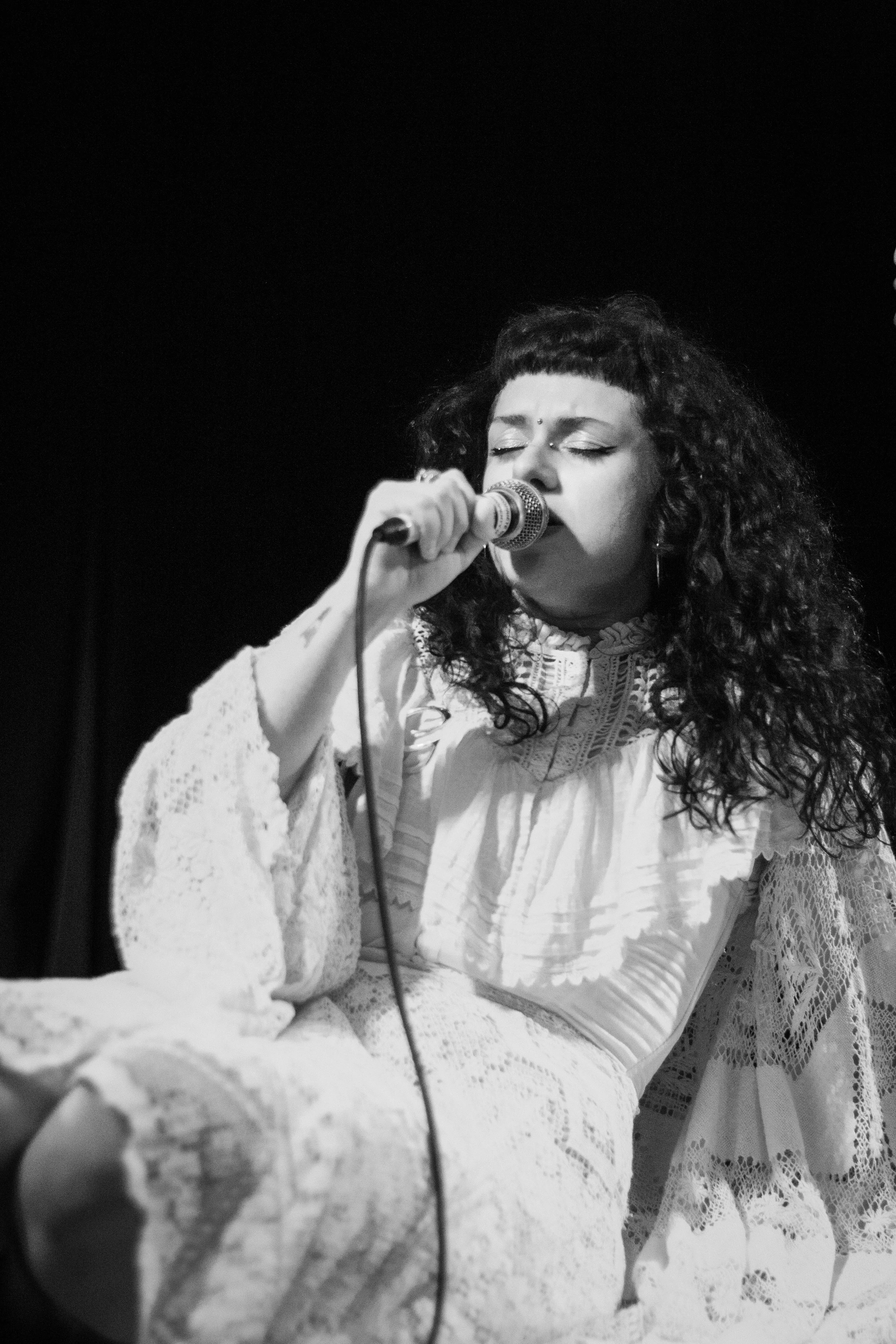 A woman with curly dark hair and closed eyes singing into a microphone. She's wearing a white lace dress and earrings, with a dark background.