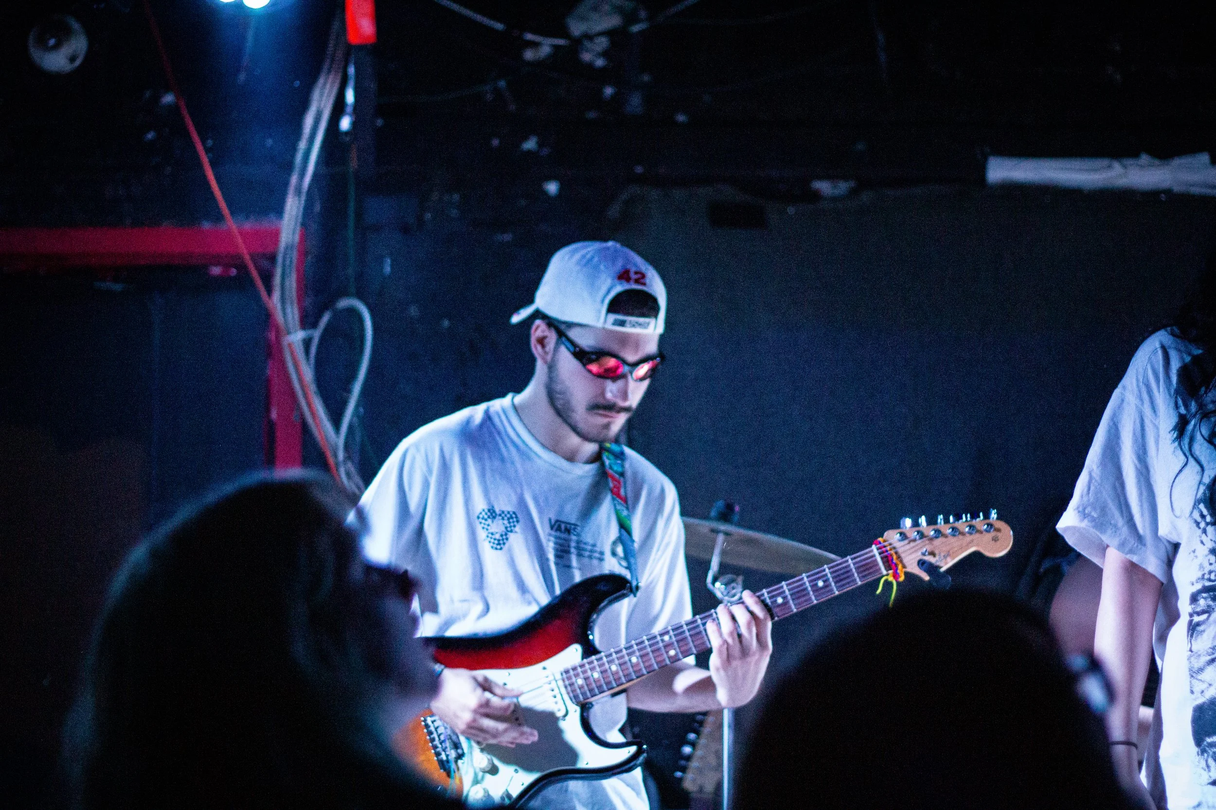 A young man wearing a white cap, black sunglasses, and a white t-shirt playing an electric guitar during a live performance in a dark room.