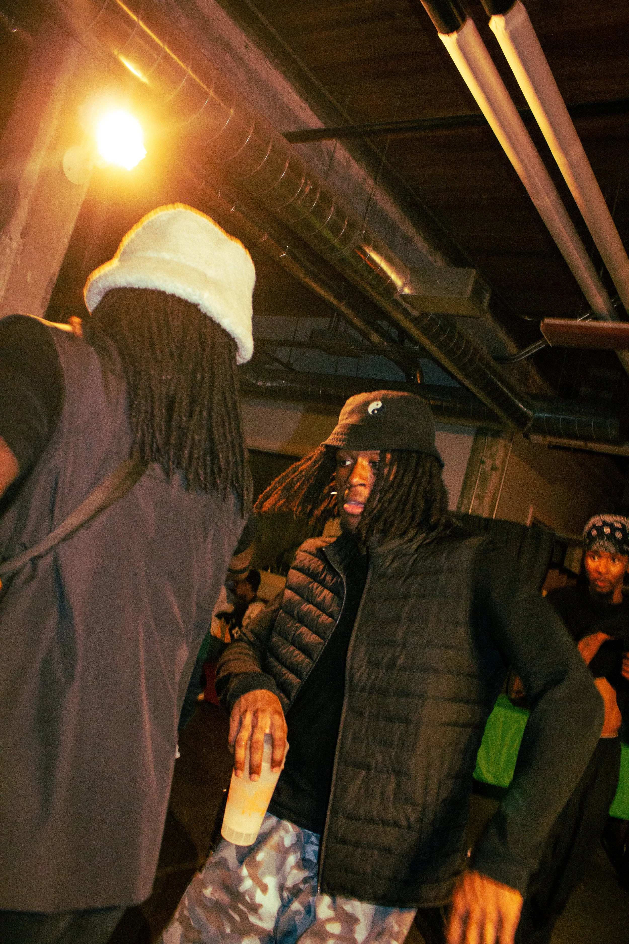 Group of people socializing indoors under industrial ceiling with black ducts and air vents, some wearing winter jackets, one person holding a phone, and a person in the background wearing a green jacket.