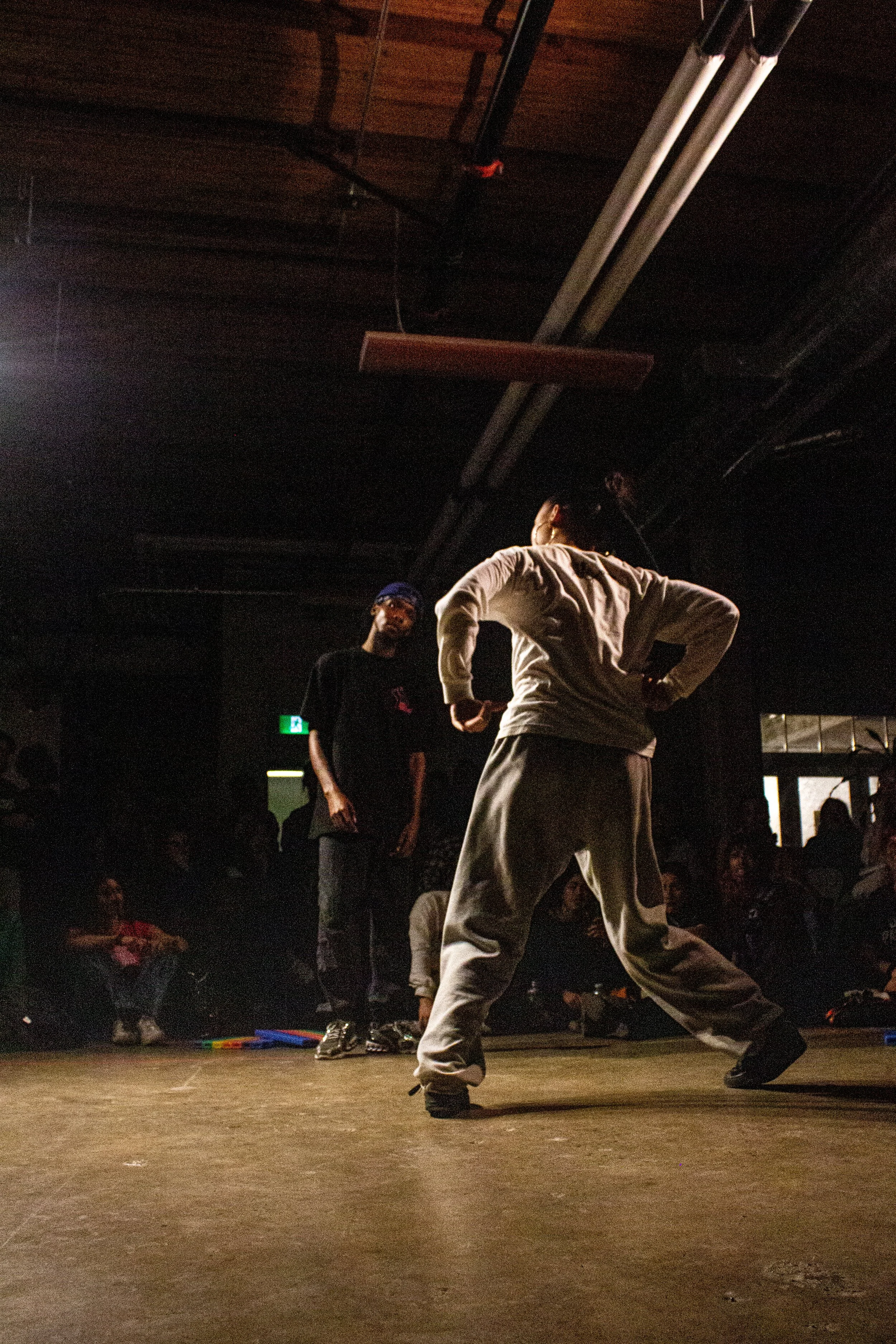 A performer on stage facing away from the camera, wearing a white outfit and black shoes, with a person in the background wearing a black shirt, black cap, and glasses, standing against a dark backdrop with ceiling lights and exposed pipes visible.