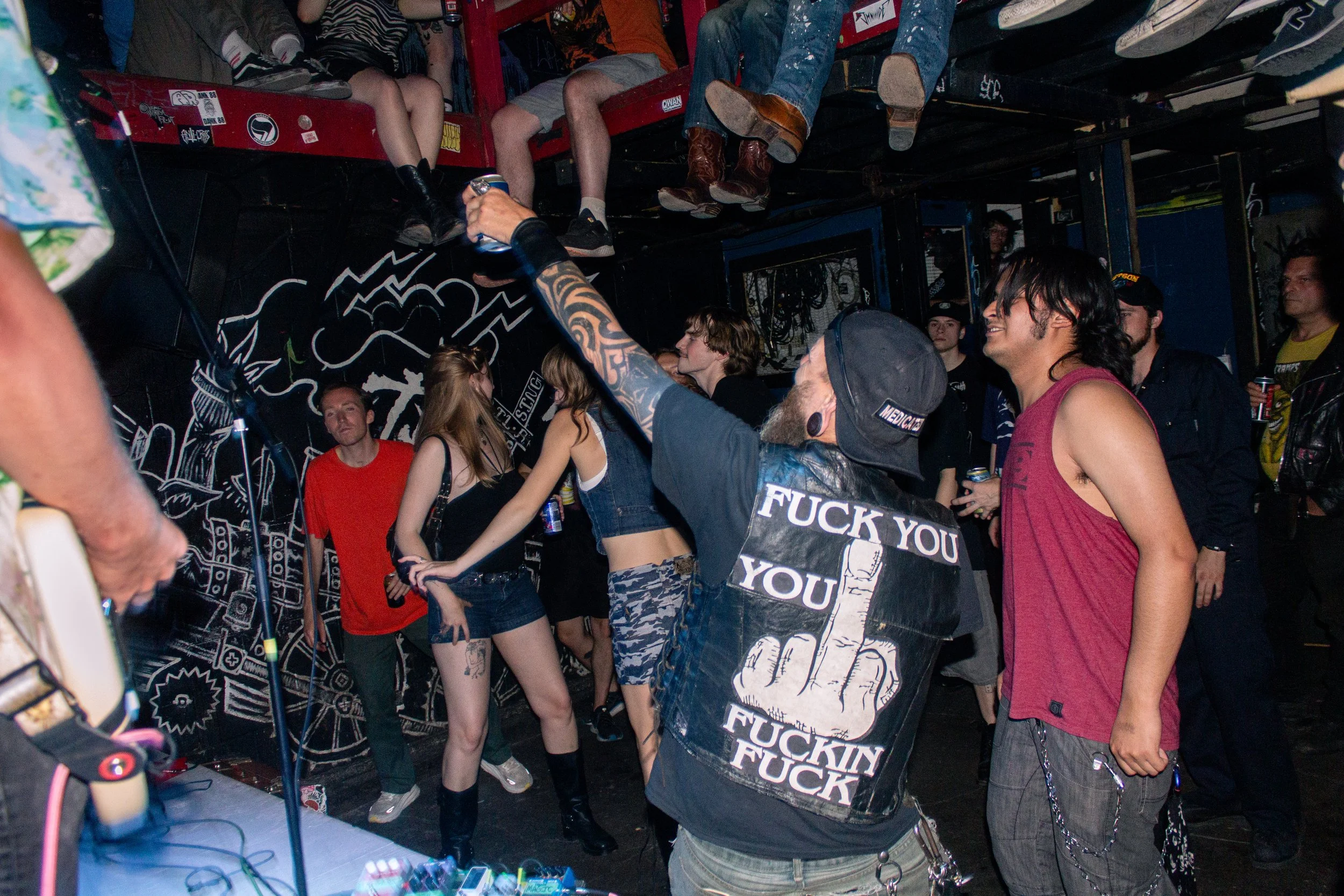 A group of young people gathered in a dimly lit venue with some people sitting on a raised platform and others standing, socializing and enjoying music. They are dressed in casual and punk-inspired clothing, with some wearing tattoos and accessories.