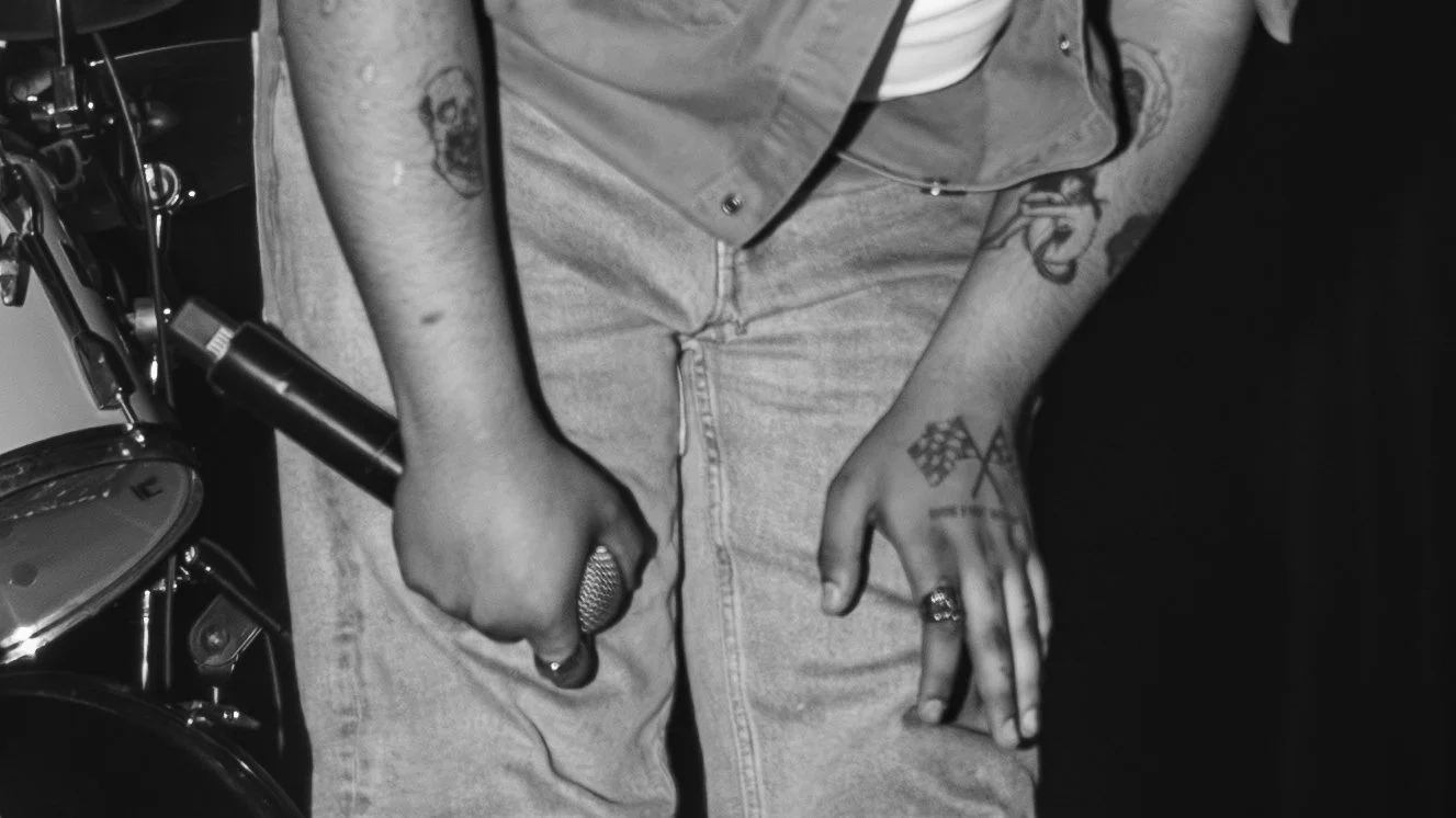 Close-up of a person sitting, showing their tattoos, rings, and hand resting on their lap, with musical equipment nearby.