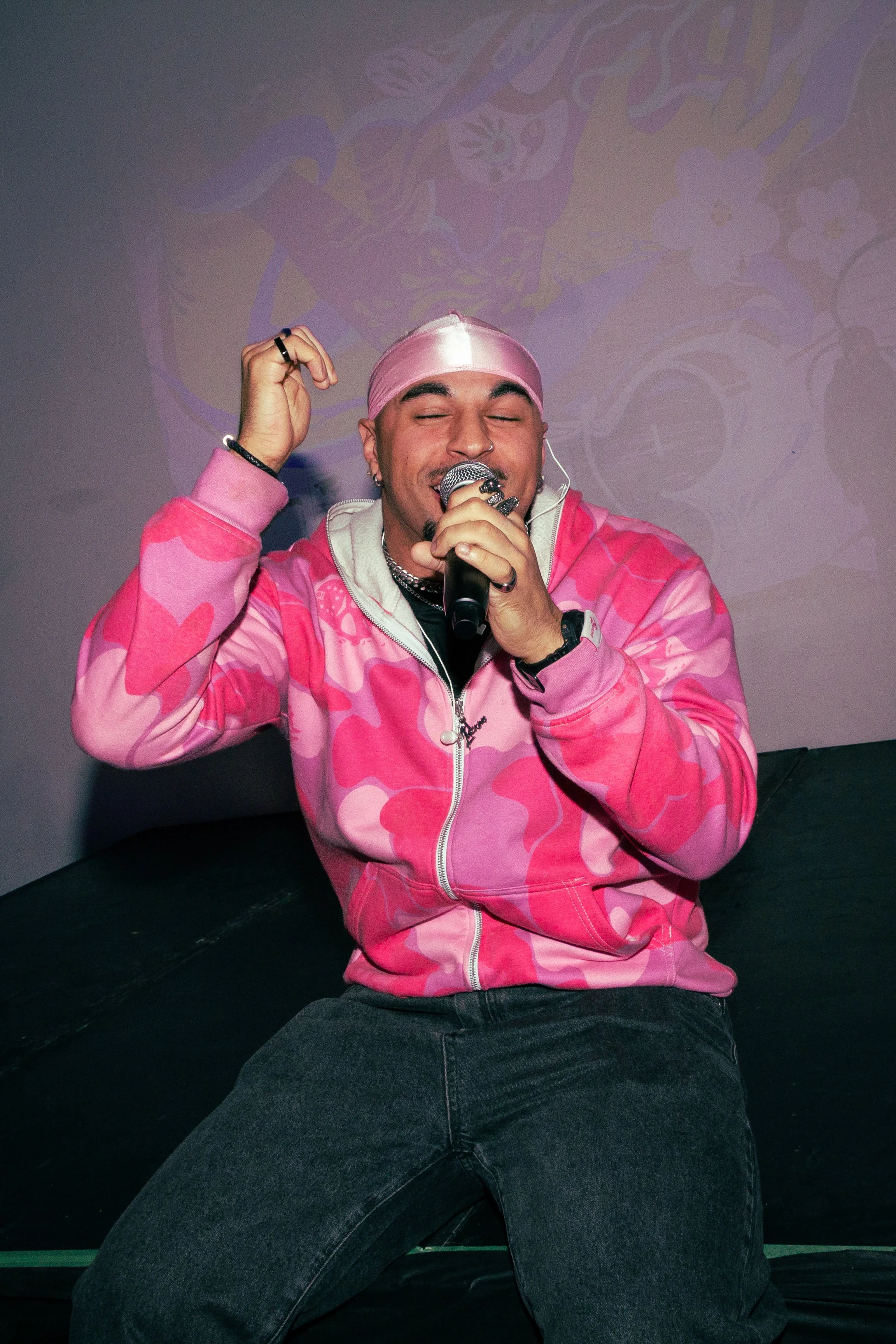 A young man wearing a pink bandana and a pink camouflage-patterned hoodie, smiling with his eyes closed, holding a microphone with his right hand, sitting on a dark surface with a purple wall background.