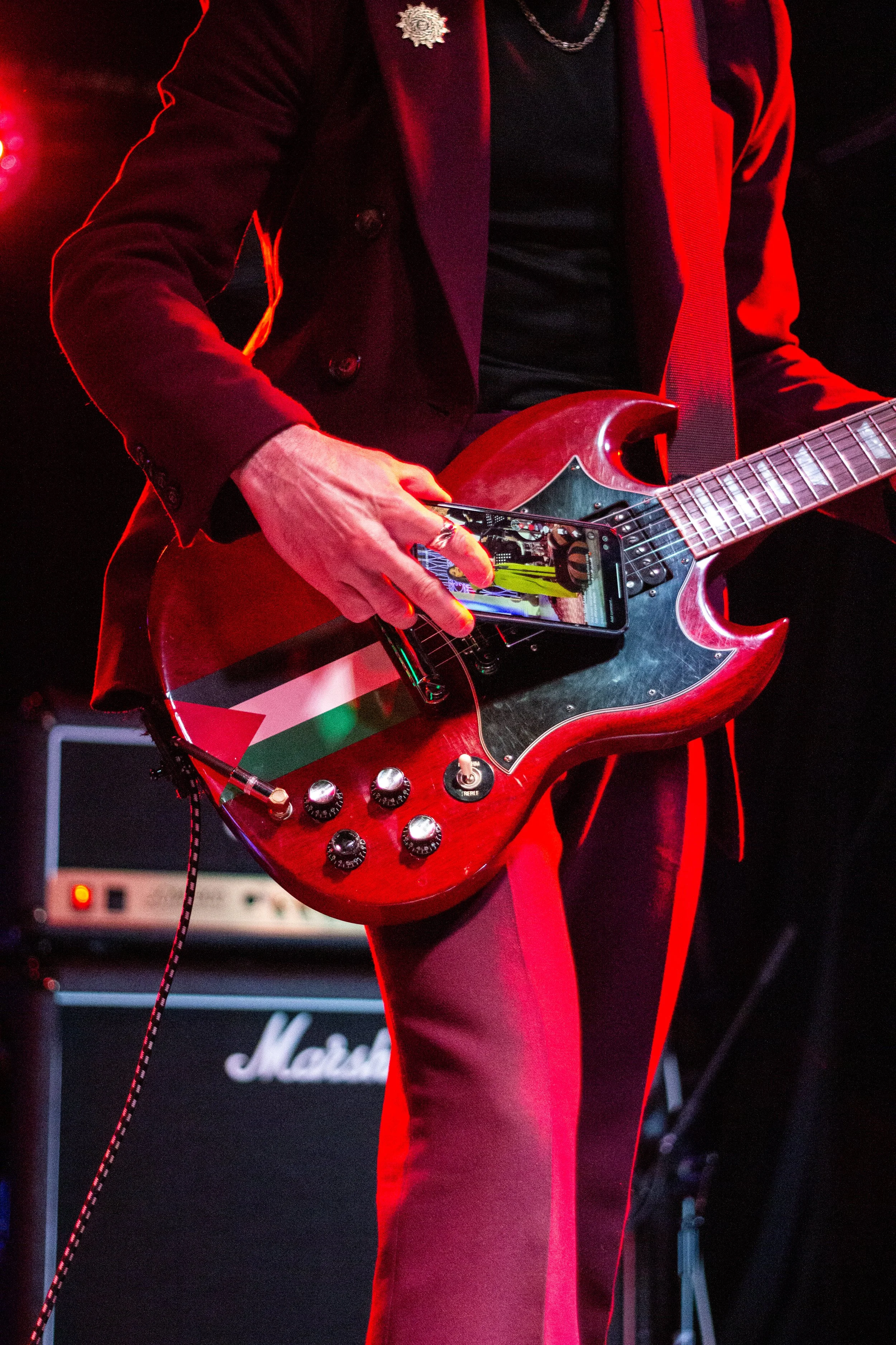 A person wearing a black jacket with a silver flower brooch, a black shirt, and red pants, is holding a guitar with a colorful sticker on it. The person appears to be on stage with a Marshall amplifier and other musical equipment in the background, a
