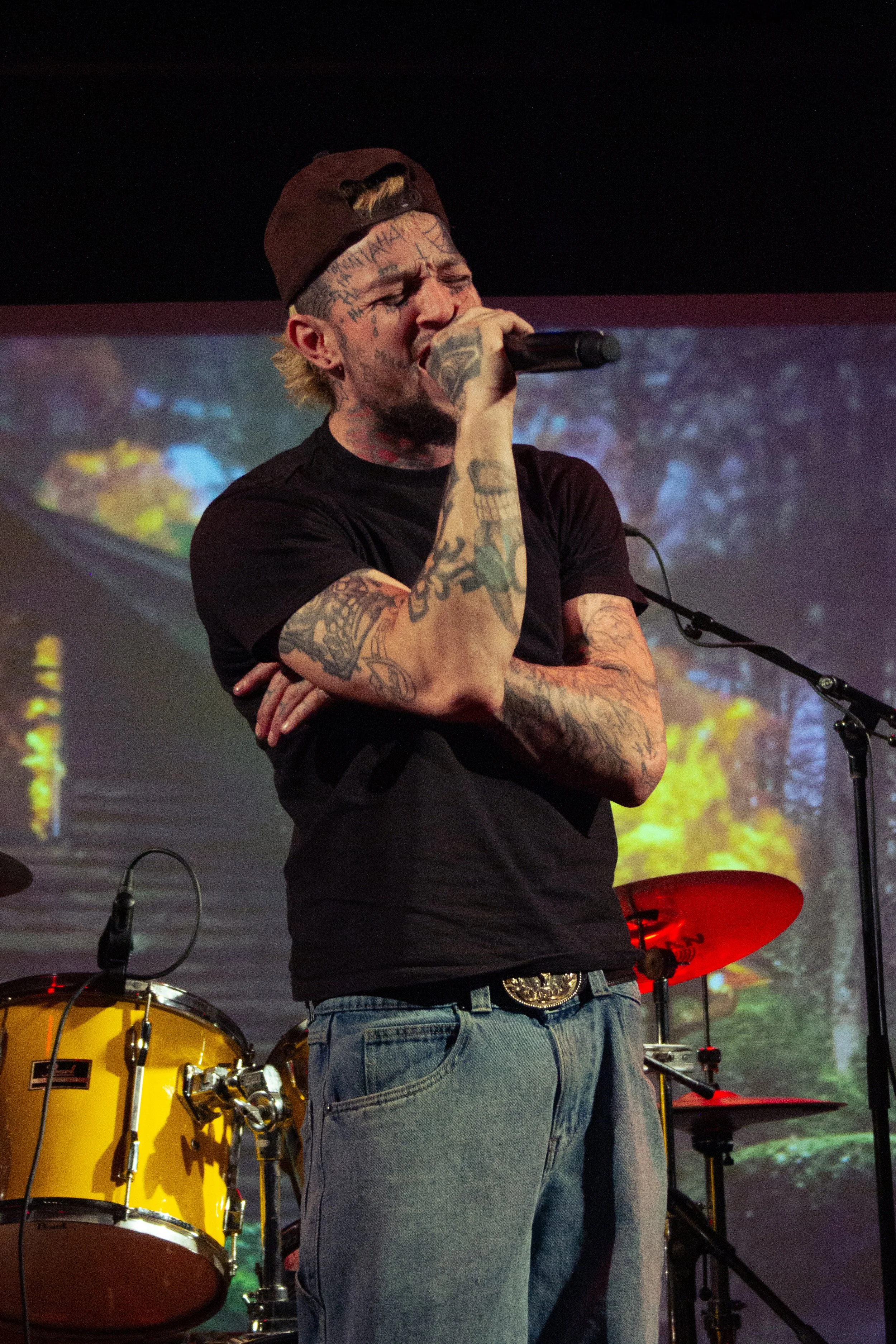 A man with tattoos singing into a microphone on stage, with yellow drums and a digital background projection behind him.