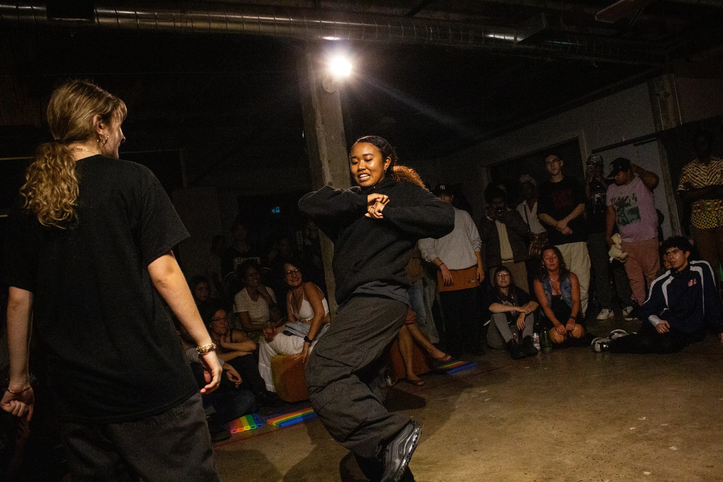 A woman with curly hair dancing indoors at a crowded event while an audience watches. There is a rainbow pride flag on the floor.