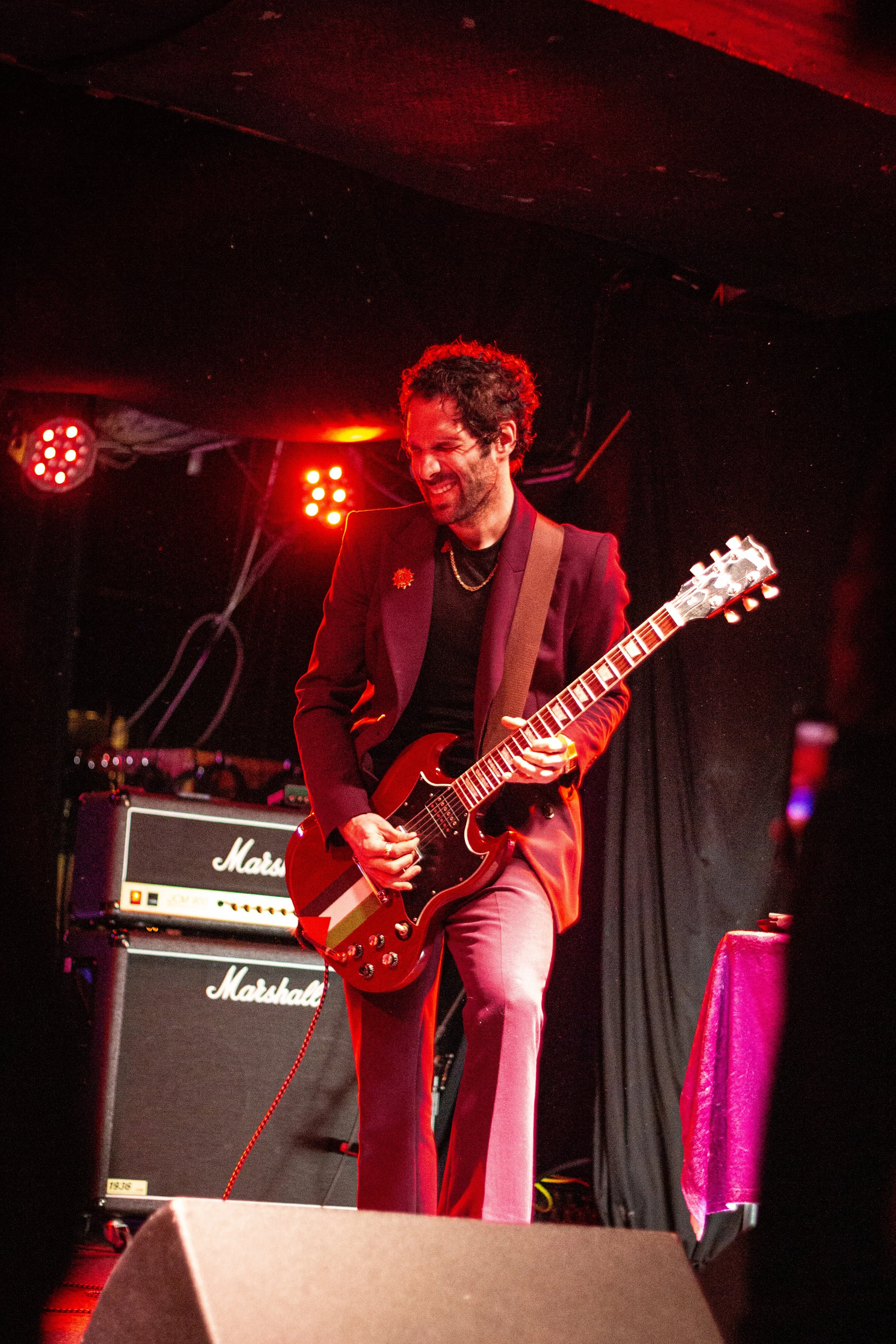 A musician playing an electric guitar on stage, with Marshall amplifiers in the background, lit by red stage lighting, and a pink cloth covering a nearby table.