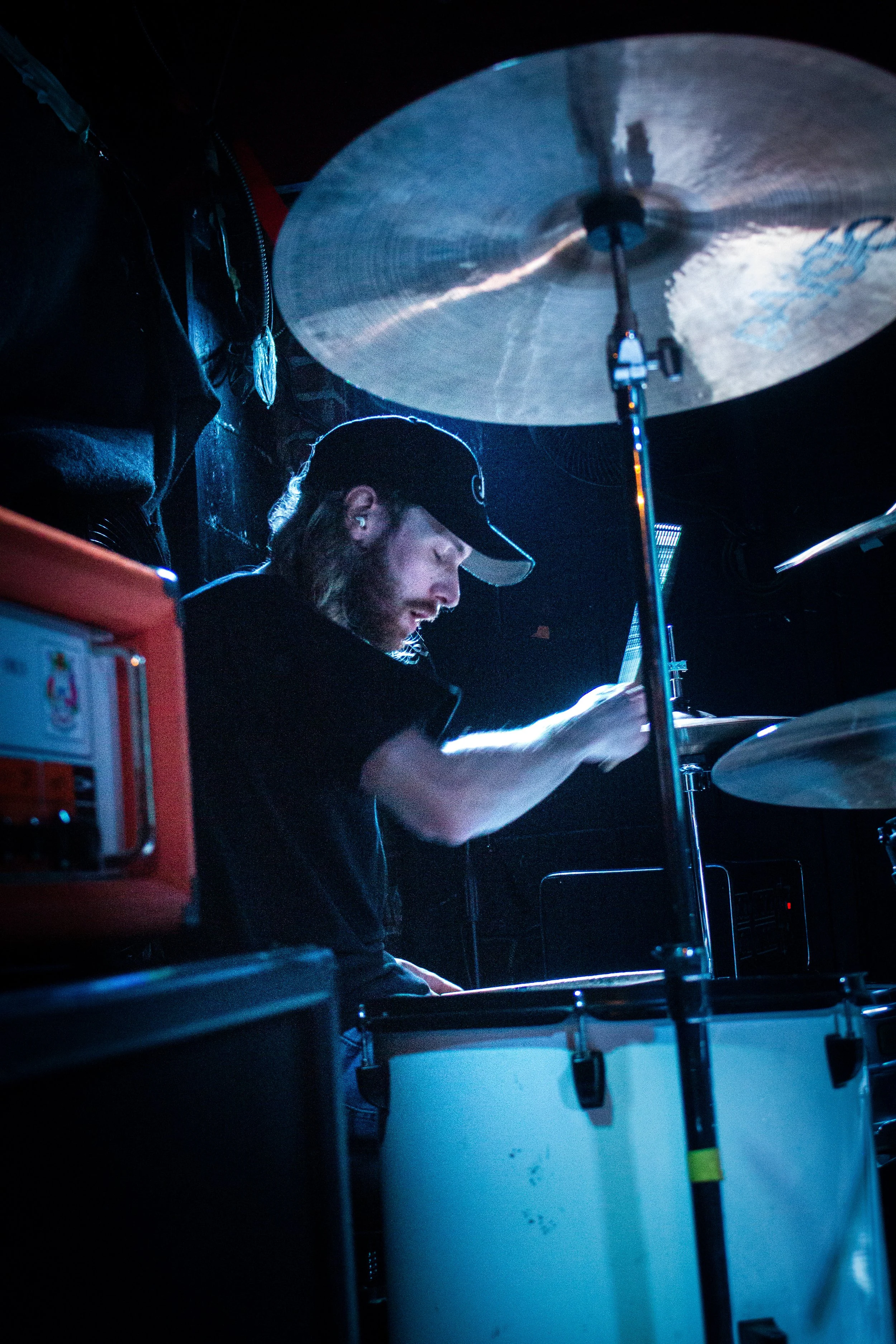 A person wearing a black cap, black jacket, and earbuds is playing drums on stage with a large cymbal and drum equipment visible in the background.