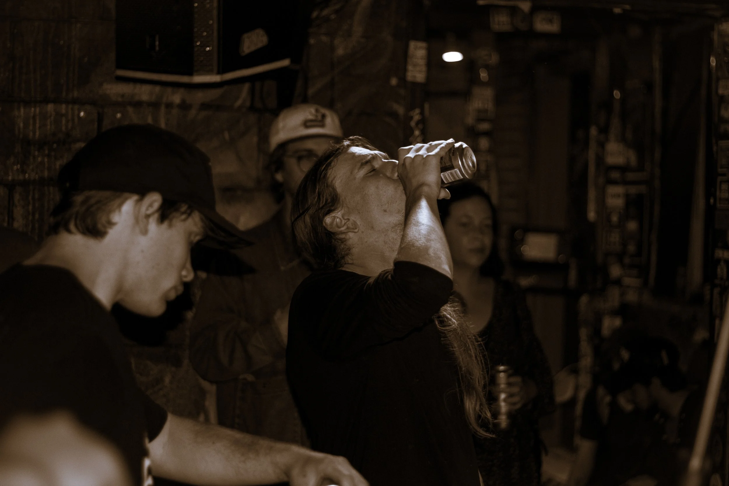 A woman drinking from a can in a dimly lit bar or pub, with other people in the background.