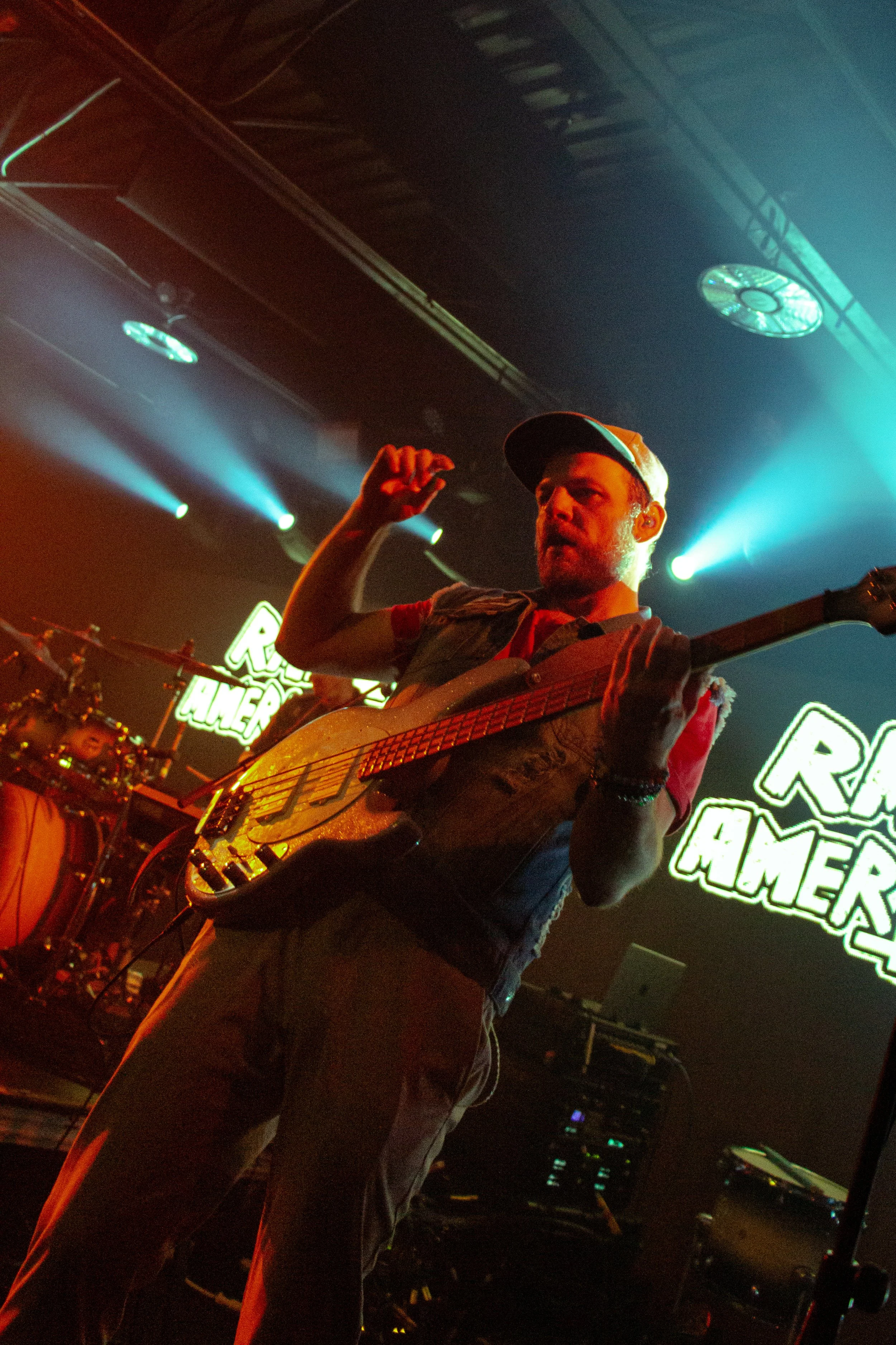 A man wearing a cap and glasses playing an electric guitar on stage under green and blue stage lights, with a drum set and illuminated signs in the background that read 'Re,' 'H,' and 'Homers'.