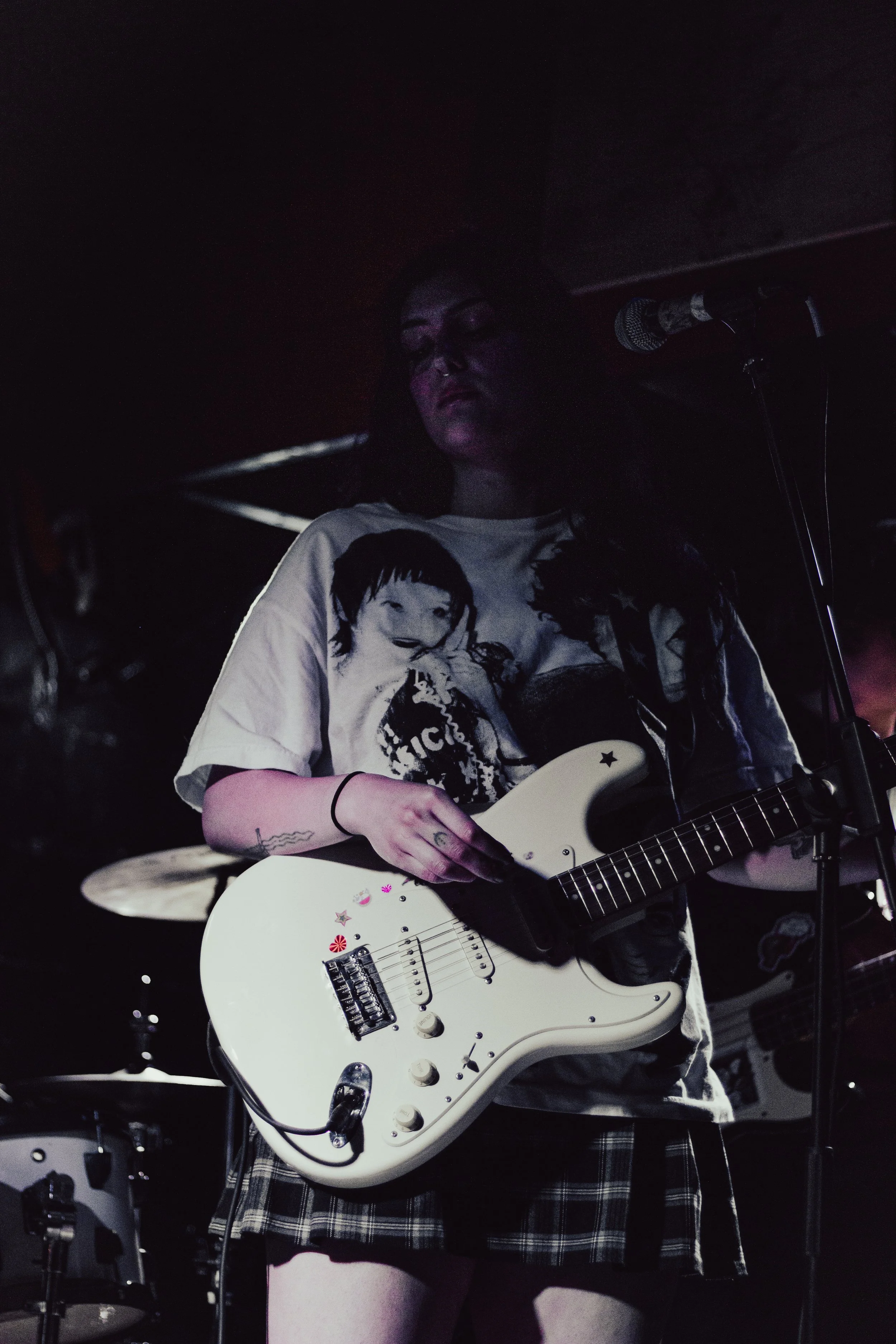 A person playing an electric guitar on stage in a dark setting, wearing a t-shirt with cartoon characters and a plaid skirt, with a microphone nearby and drums visible in the background.