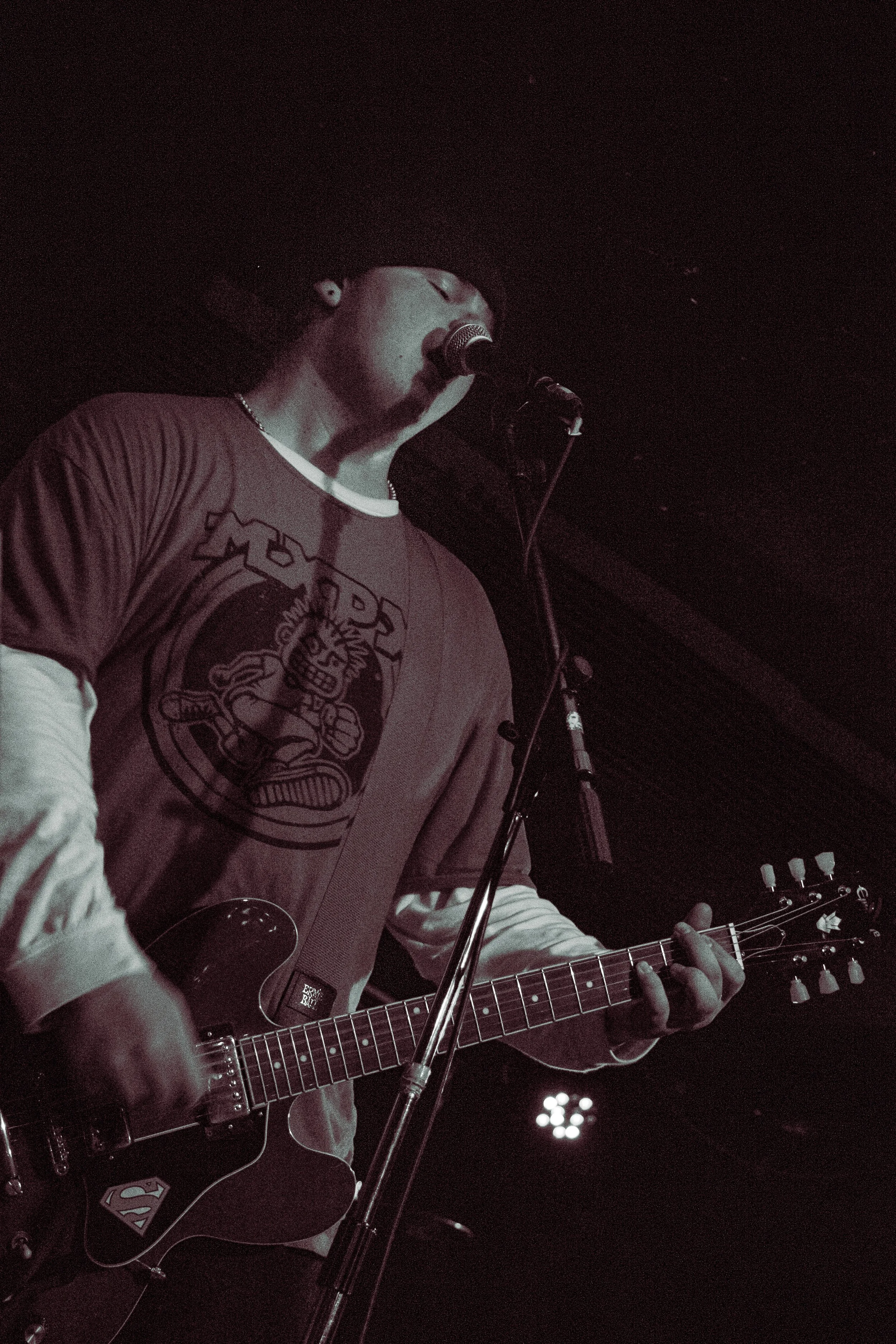 A person singing into a microphone while playing a guitar on stage, wearing a  t-shirt and a beaded necklace.