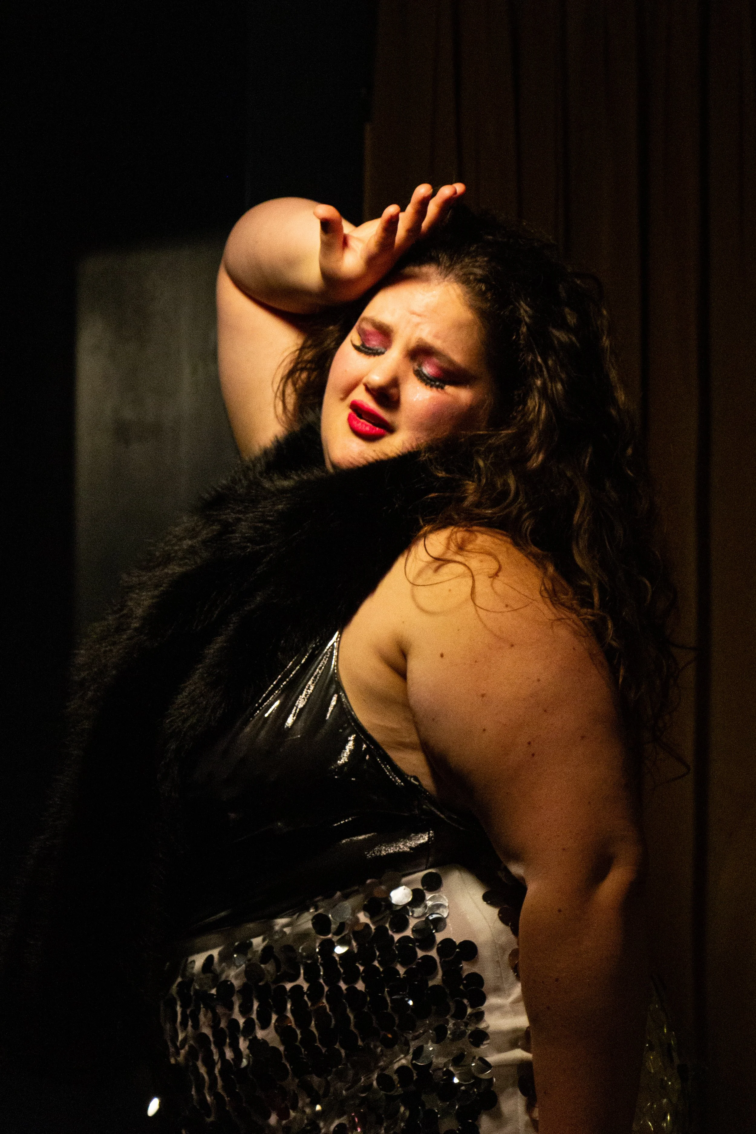 A woman with curly hair and makeup, wearing a black top with a sequin or mirror-like pattern, posing with her hand on her forehead, in a dimly lit or dark setting.