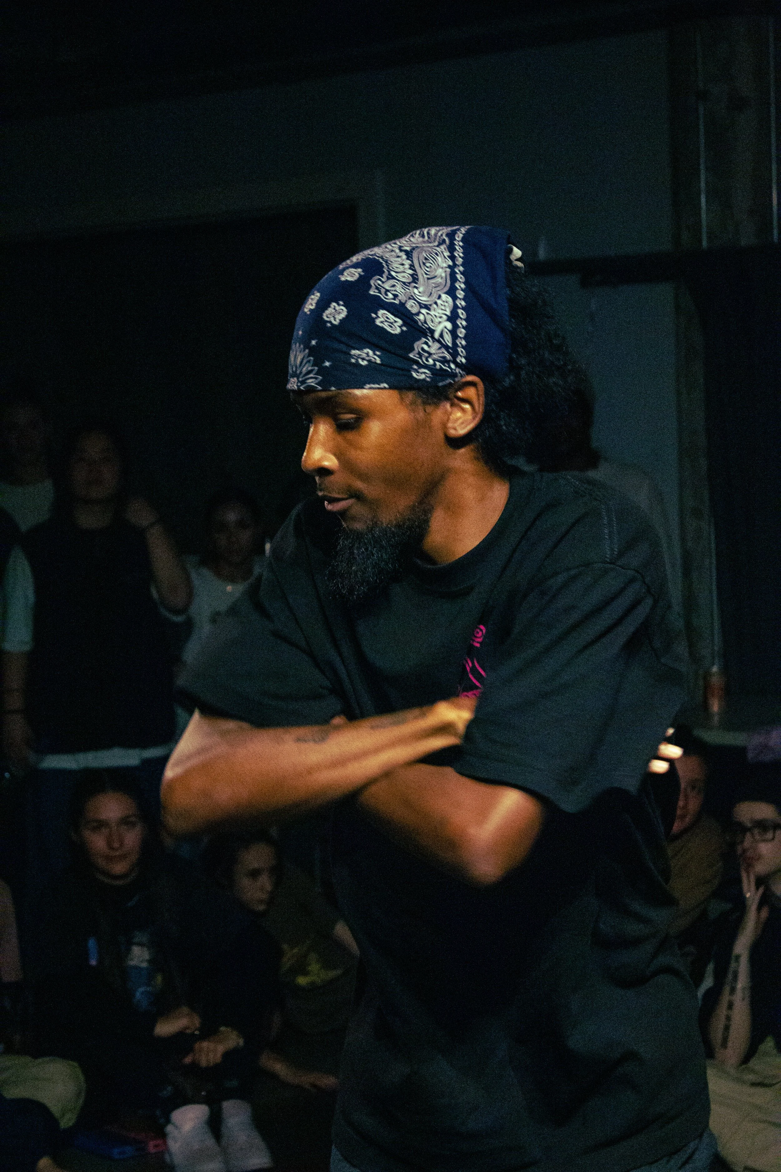 A man with a beard and curly hair wearing a blue bandana and black shirt, standing with arms crossed in front of a group of people in a dimly lit room.