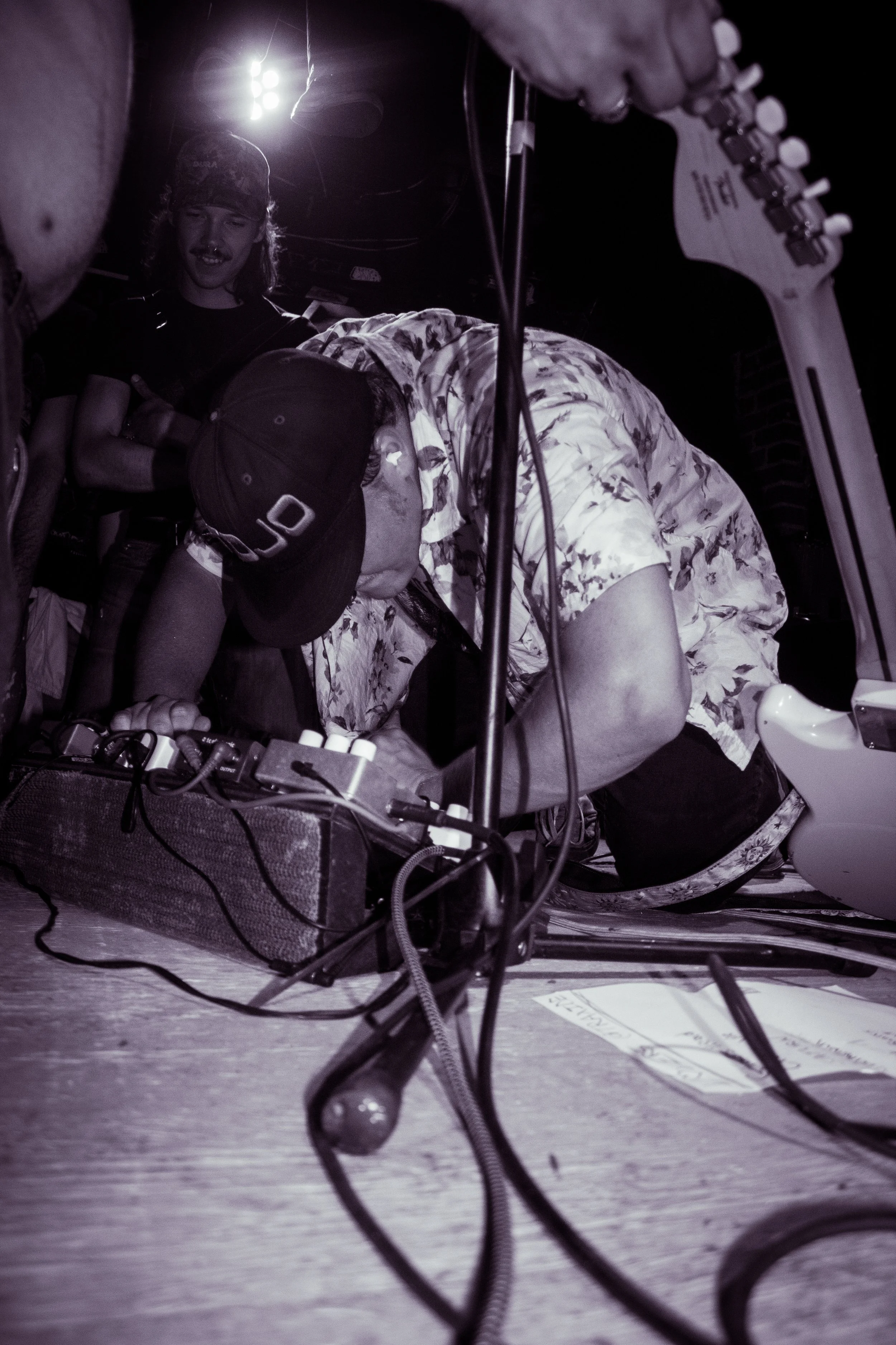 A musician wearing a floral shirt and a black cap is playing an electric guitar on a stage, with another person standing nearby smiling. There is electronic music equipment on the floor in front of them, and stage lights in the background.