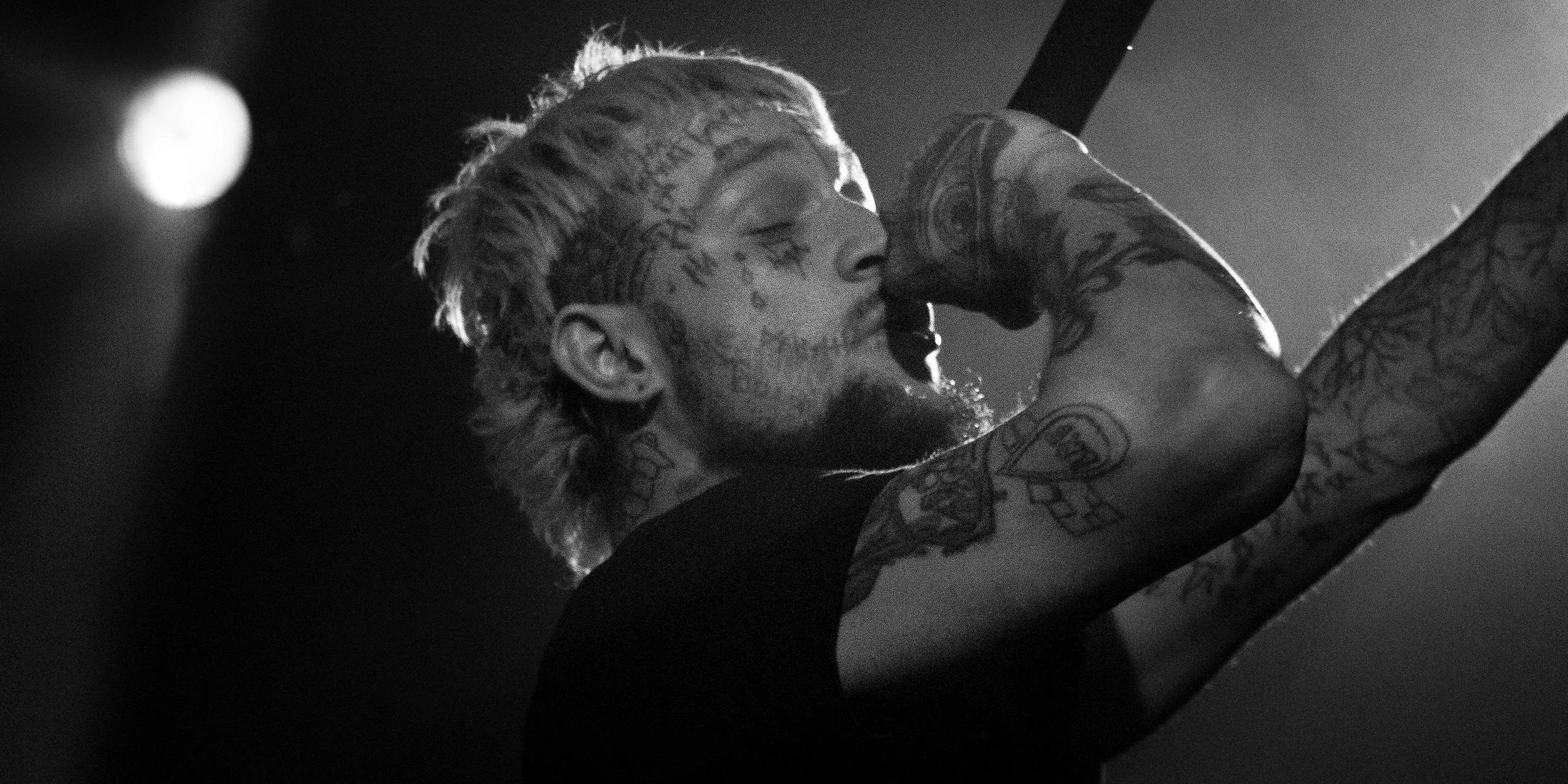 Black and white photo of a tattooed man with light-colored hair, tattoos on his face and arms, licking his wrist with his eyes closed, in a dramatic lighting environment.