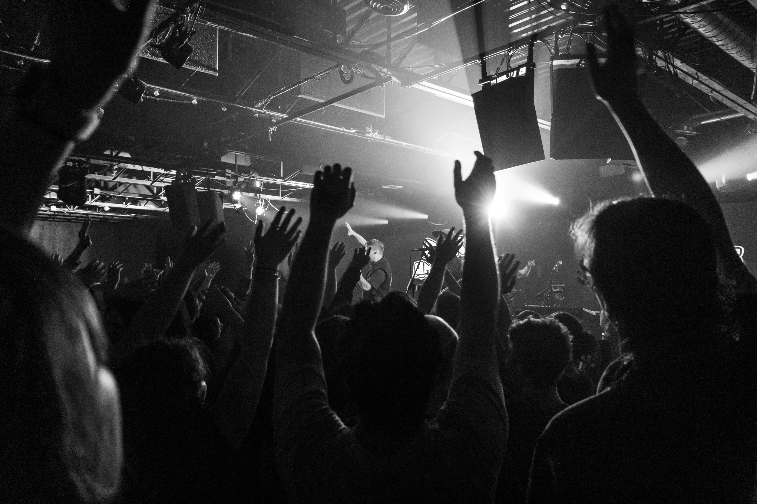 Black and white photo of a concert or live music event with an audience raising their hands, stage lighting illuminating the space, and performers visible in the background.