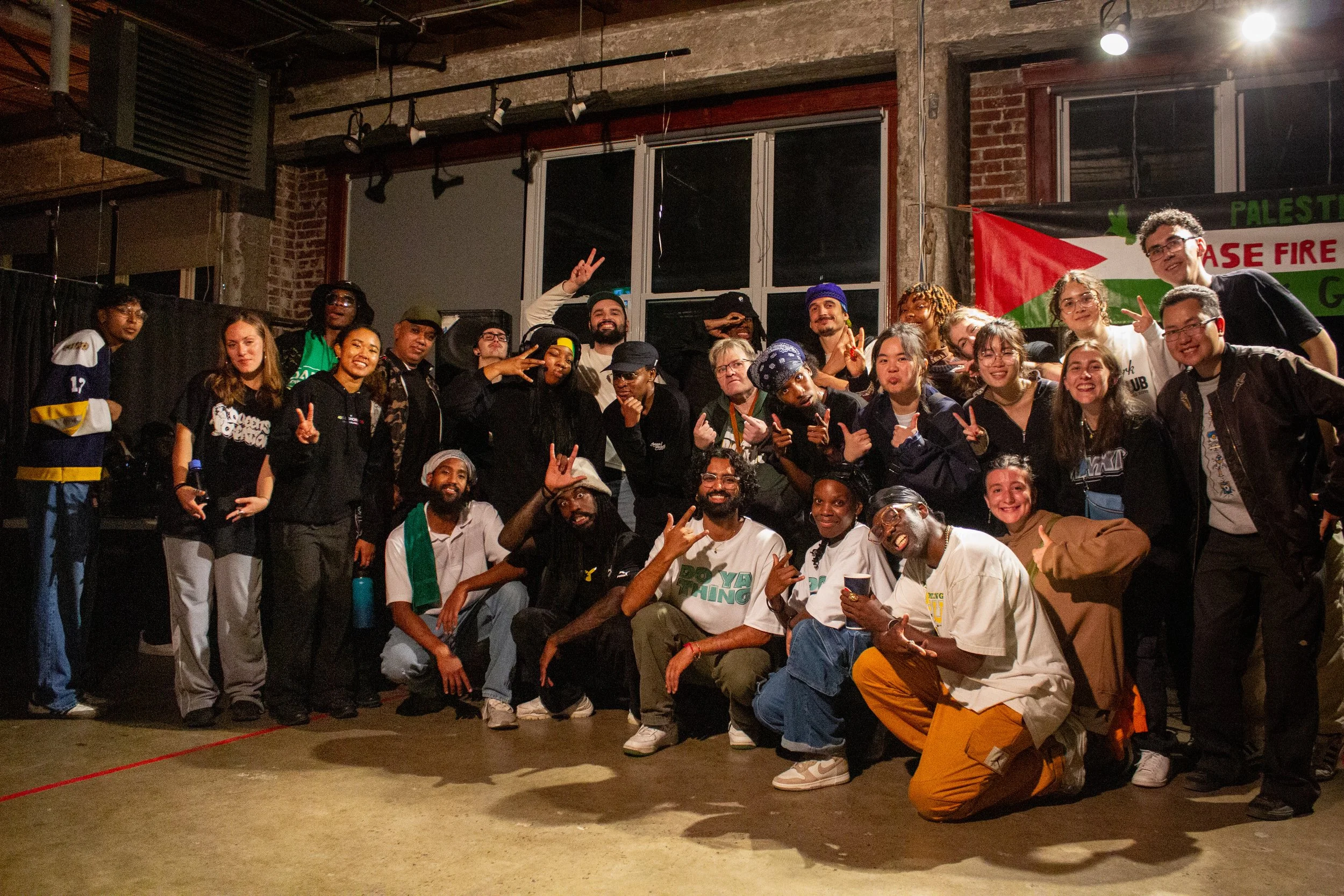 Group of diverse people posing for a photo in an indoor space with brick walls and large windows. Some are making peace signs and smiling, with a banner in the background that reads 'Palestine' and 'Case Fire'.