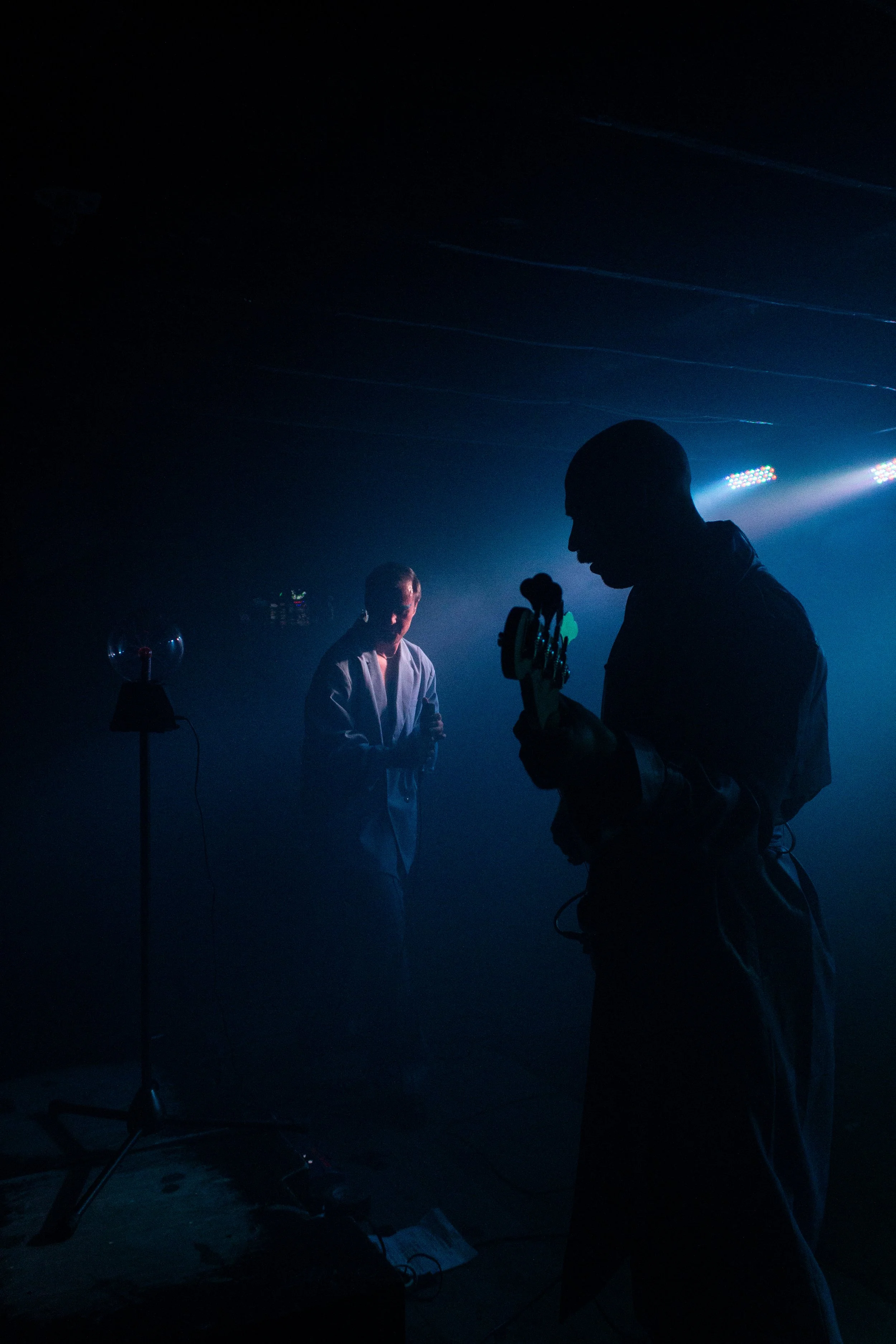 Silhouettes of two people in a dark room with stage lights, one holding a guitar and the other standing nearby.