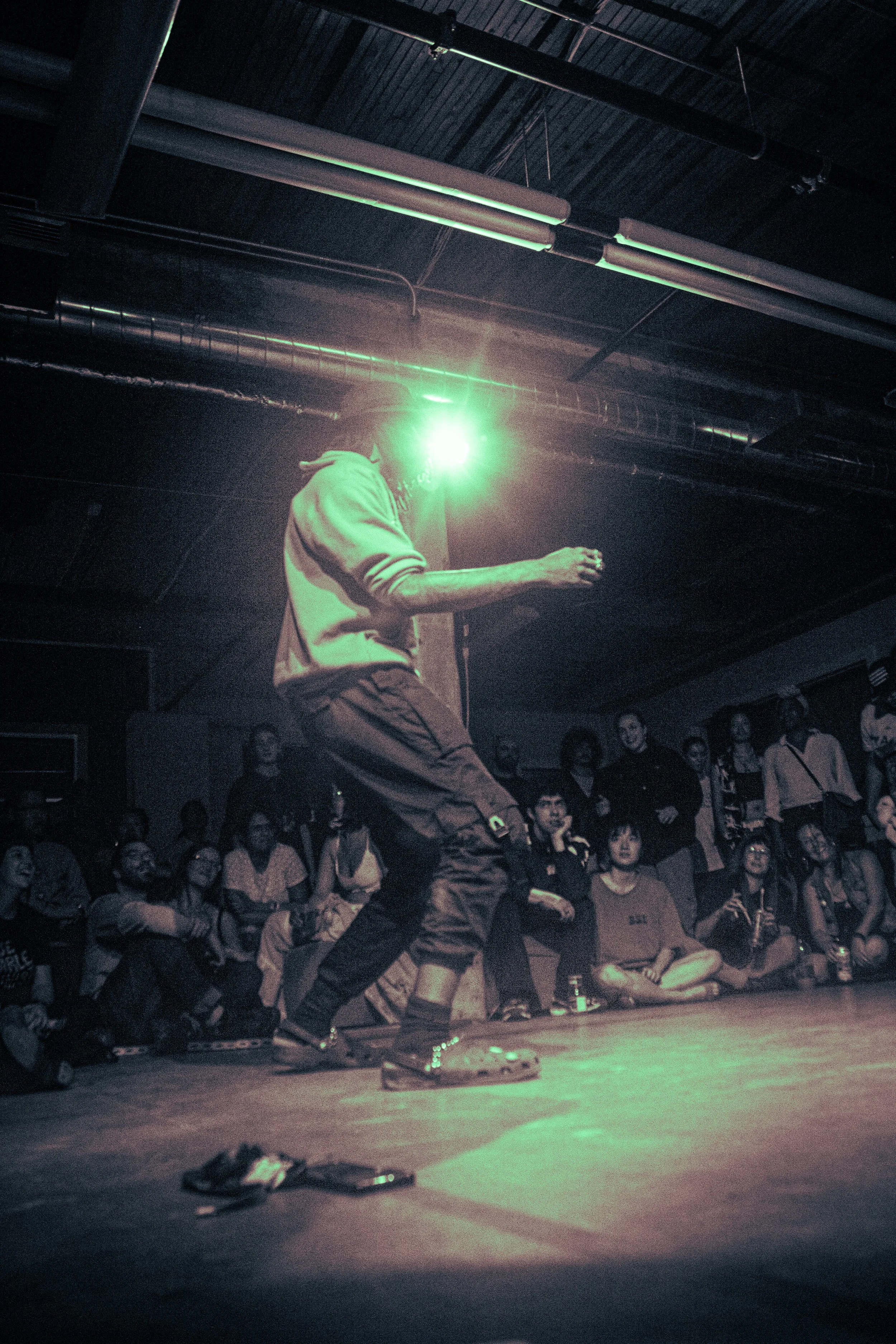 A dancer performs on a stage illuminated by green light in front of an audience in a dark room with visible ceiling pipes.