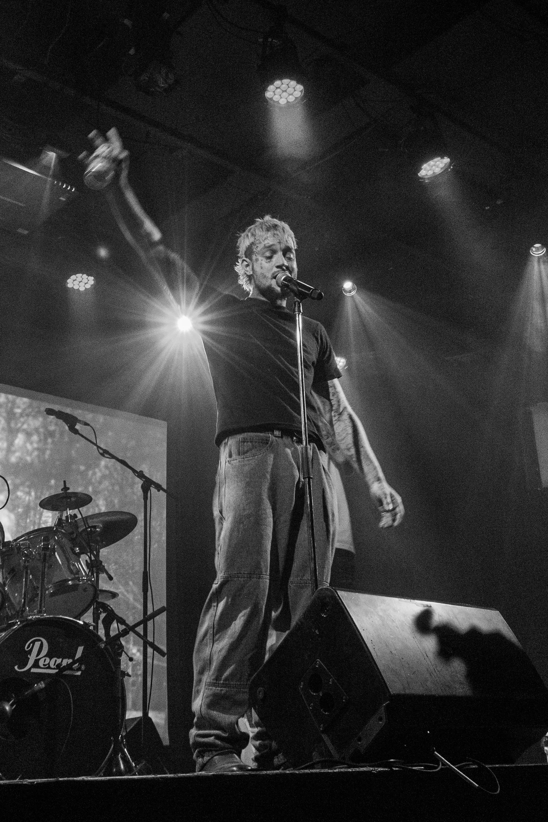 A male singer with light-colored hair singing on stage into a microphone, wearing a dark shirt and jeans. Stage lights shine behind him, and musical instruments, including a drum set, are visible in the background.