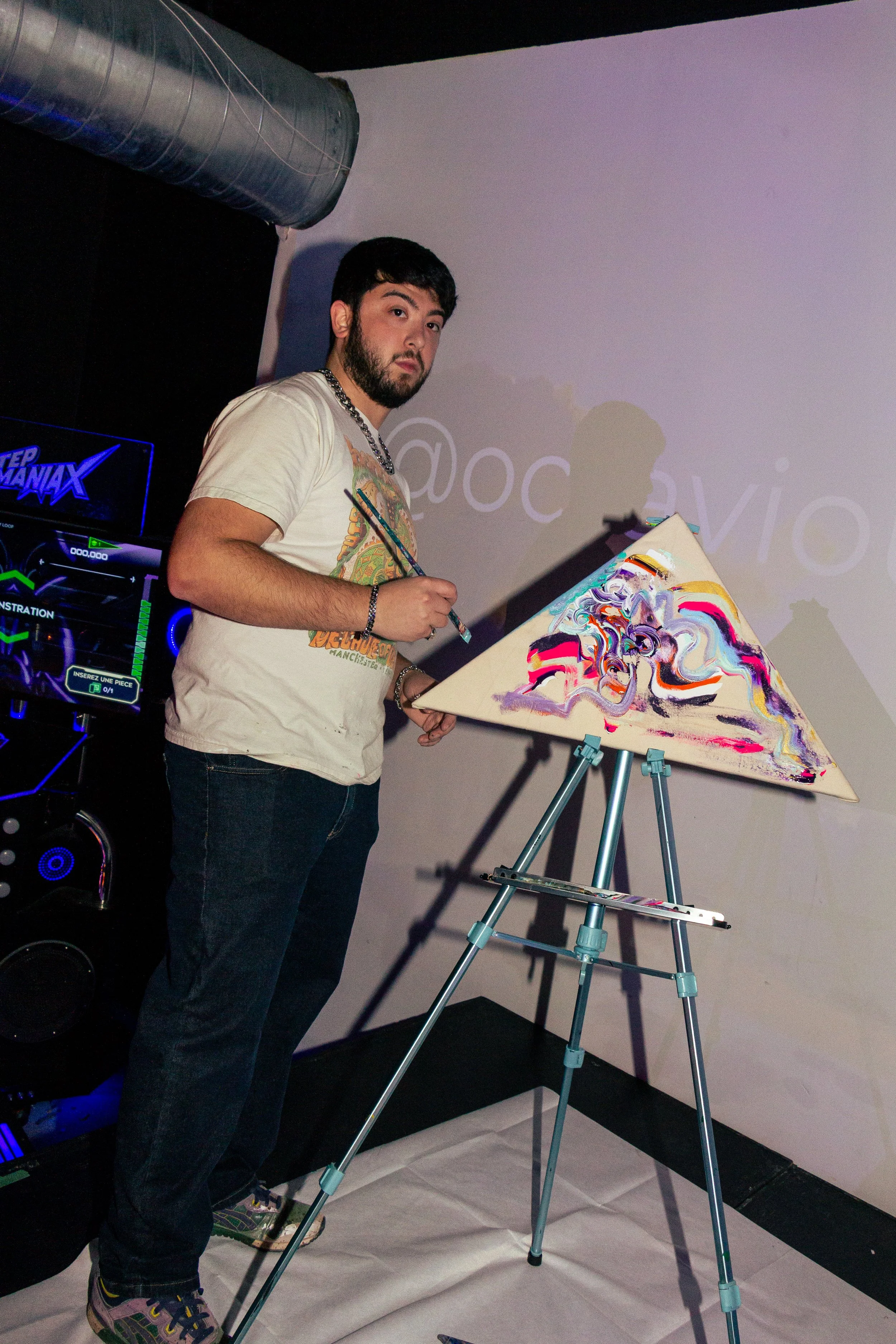 Young man standing in front of a blank wall, holding a paintbrush and a canvas with abstract colorful artwork on an easel, wearing a beige T-shirt with a graphic design, jeans, and colorful sneakers. There is a large speaker and a monitor in the back