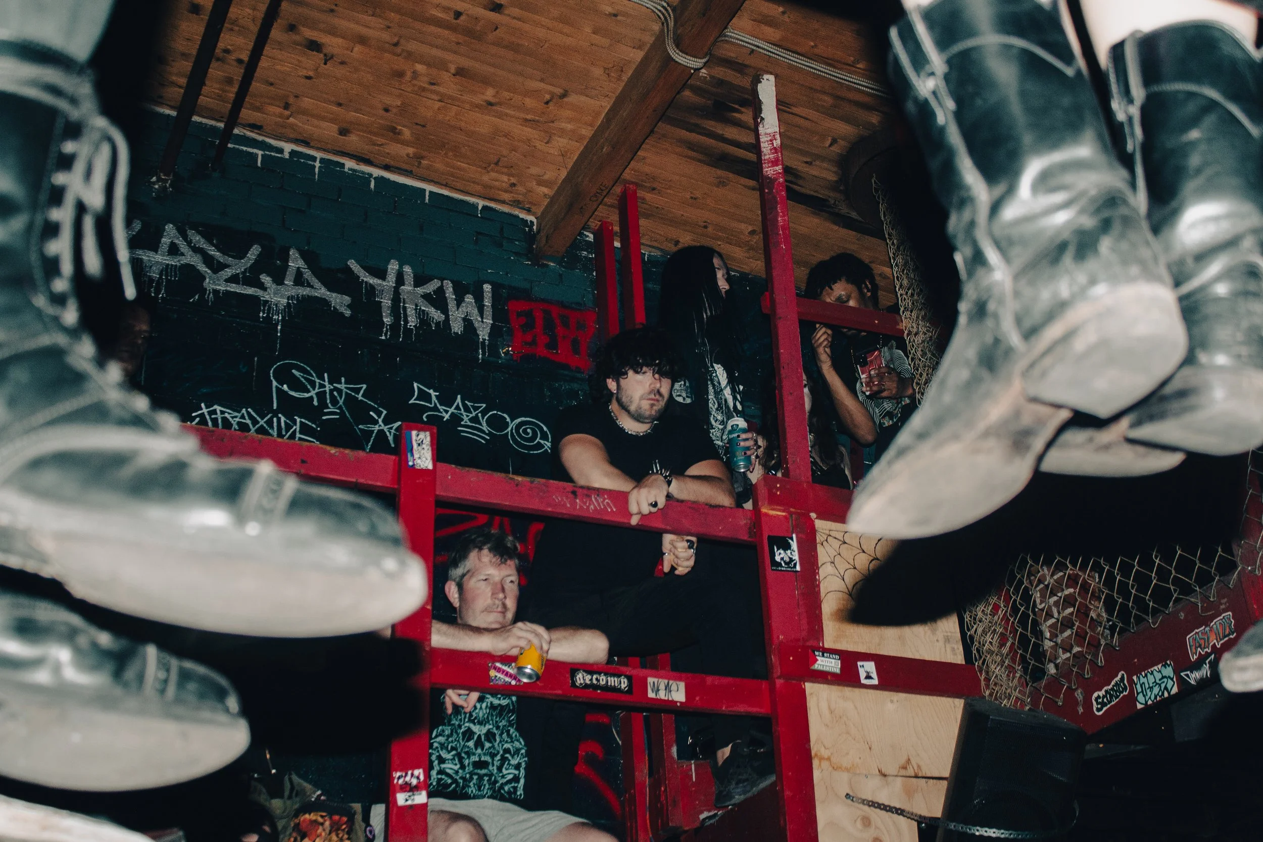 People sitting on a red metal bunk bed with graffiti on the wall behind them, viewed from underneath with a shoe in the foreground.