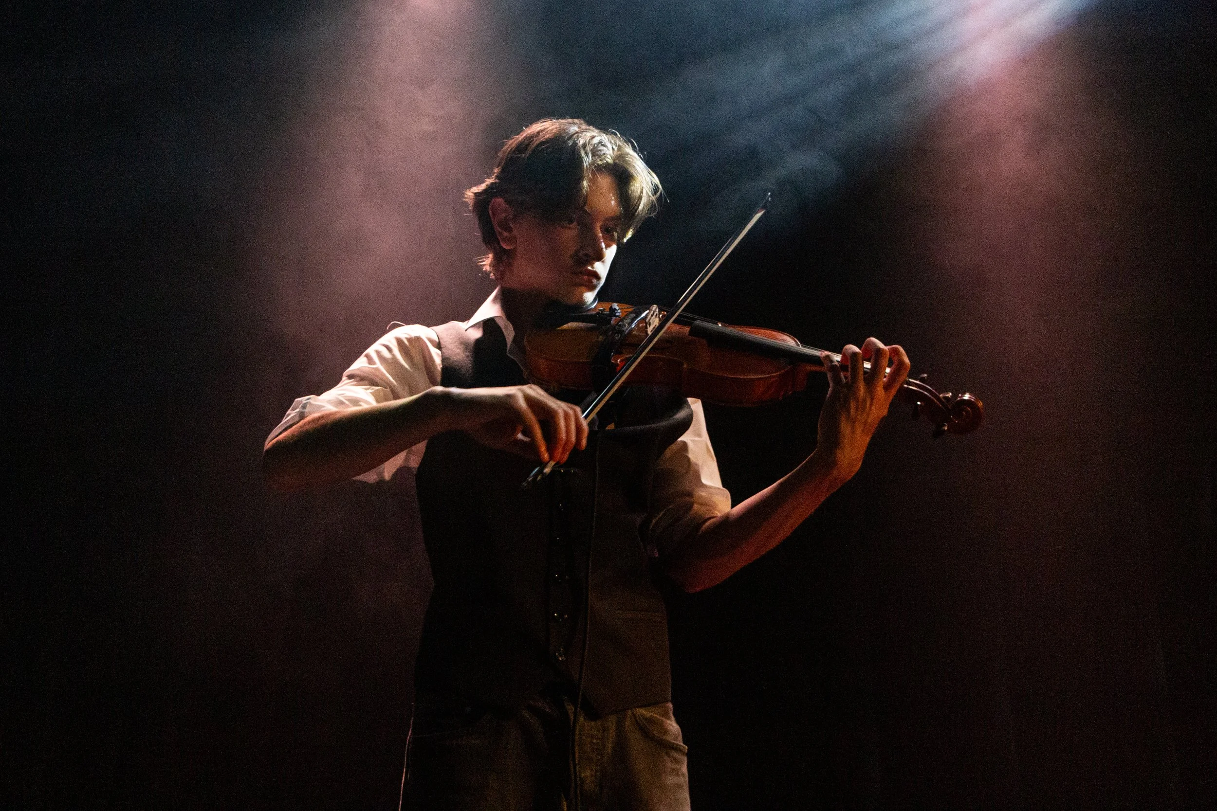 A young man playing the violin on a dimly lit stage, focused and immersed in music, with a dark background and subtle stage lighting.