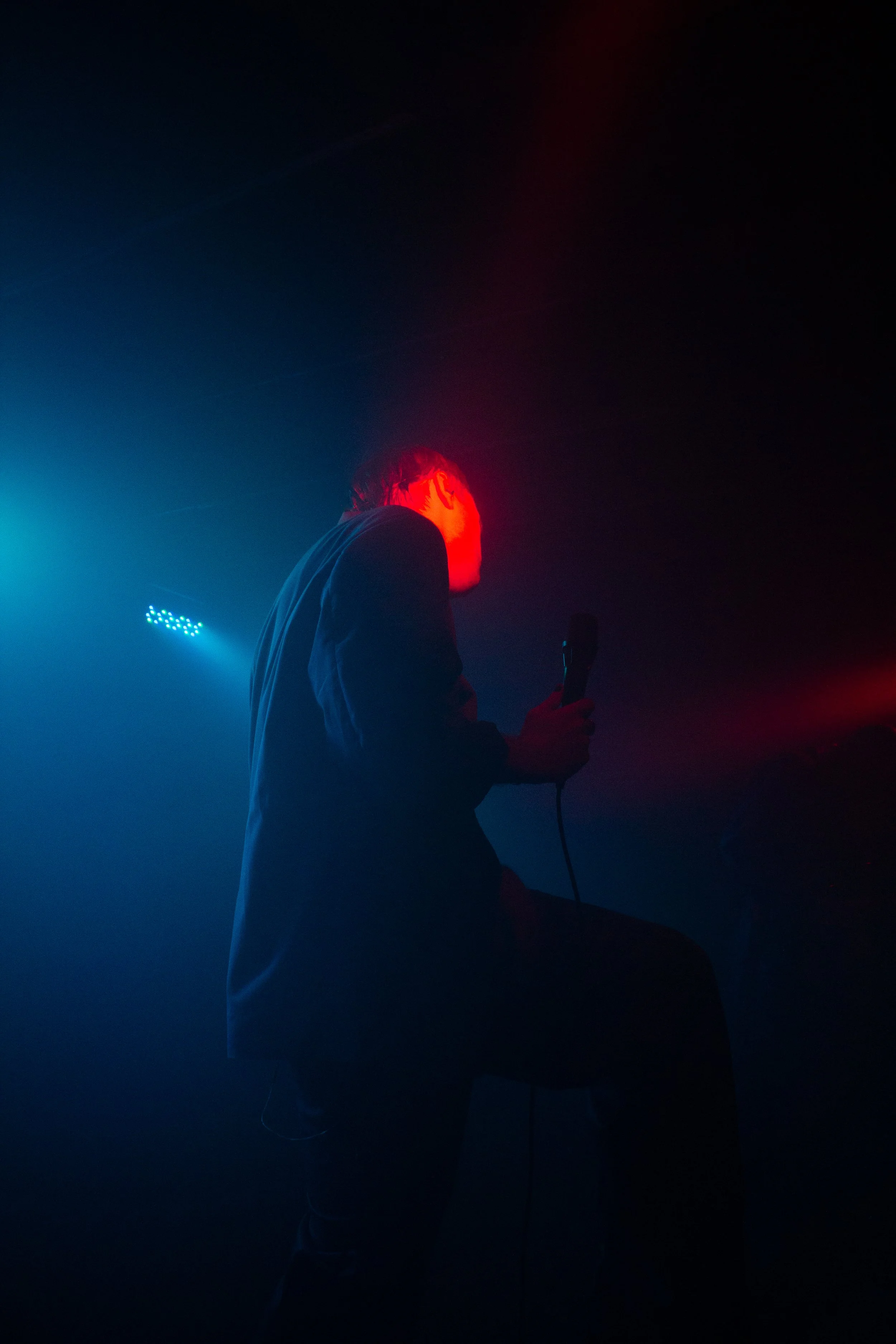A person performing on stage in front of colorful blue and red lighting, holding a microphone, with their head turned to the side