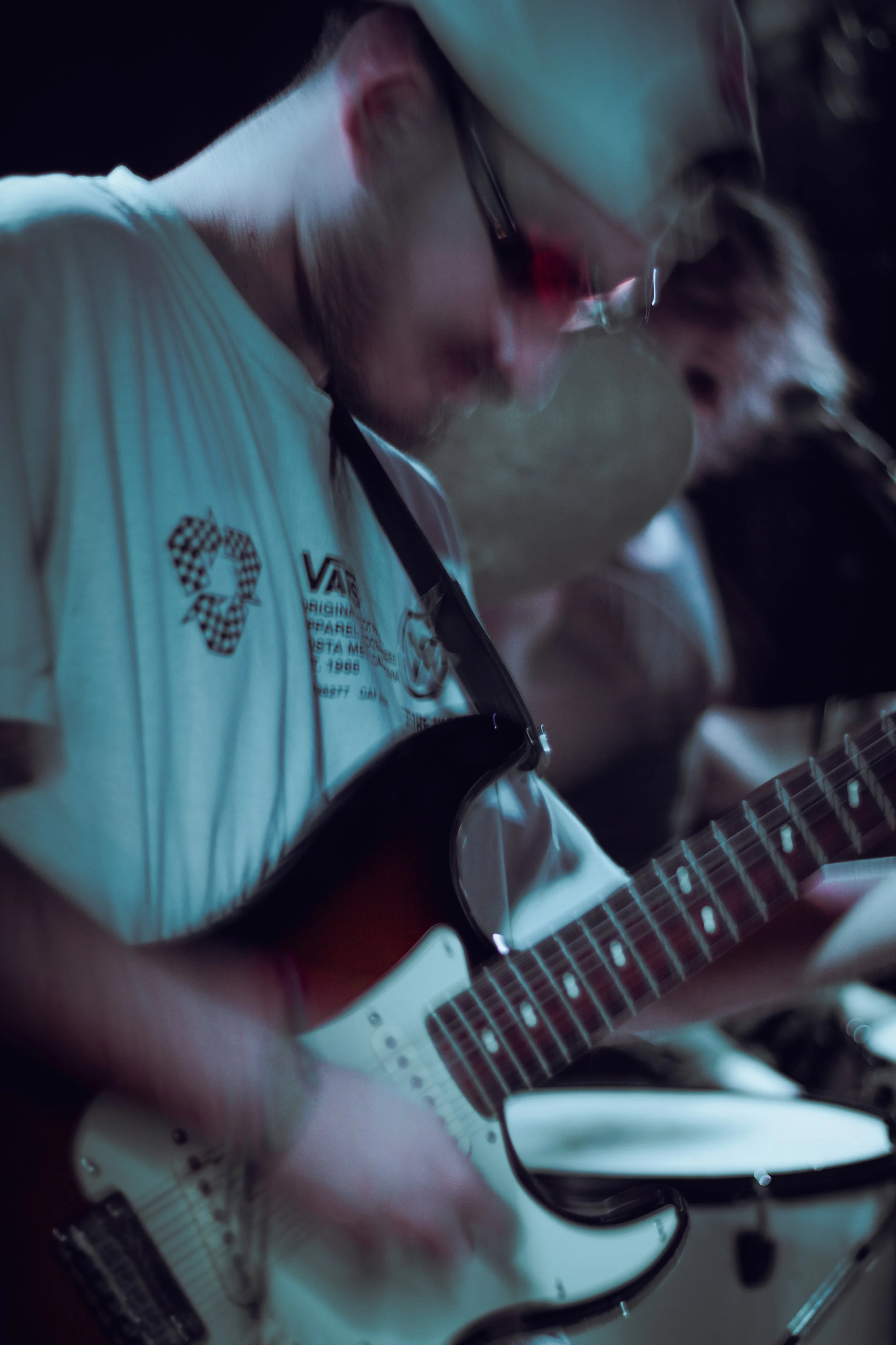 A person playing an electric guitar, wearing glasses and a white t-shirt with text and graphics, focused on the instrument in a dimly lit setting.