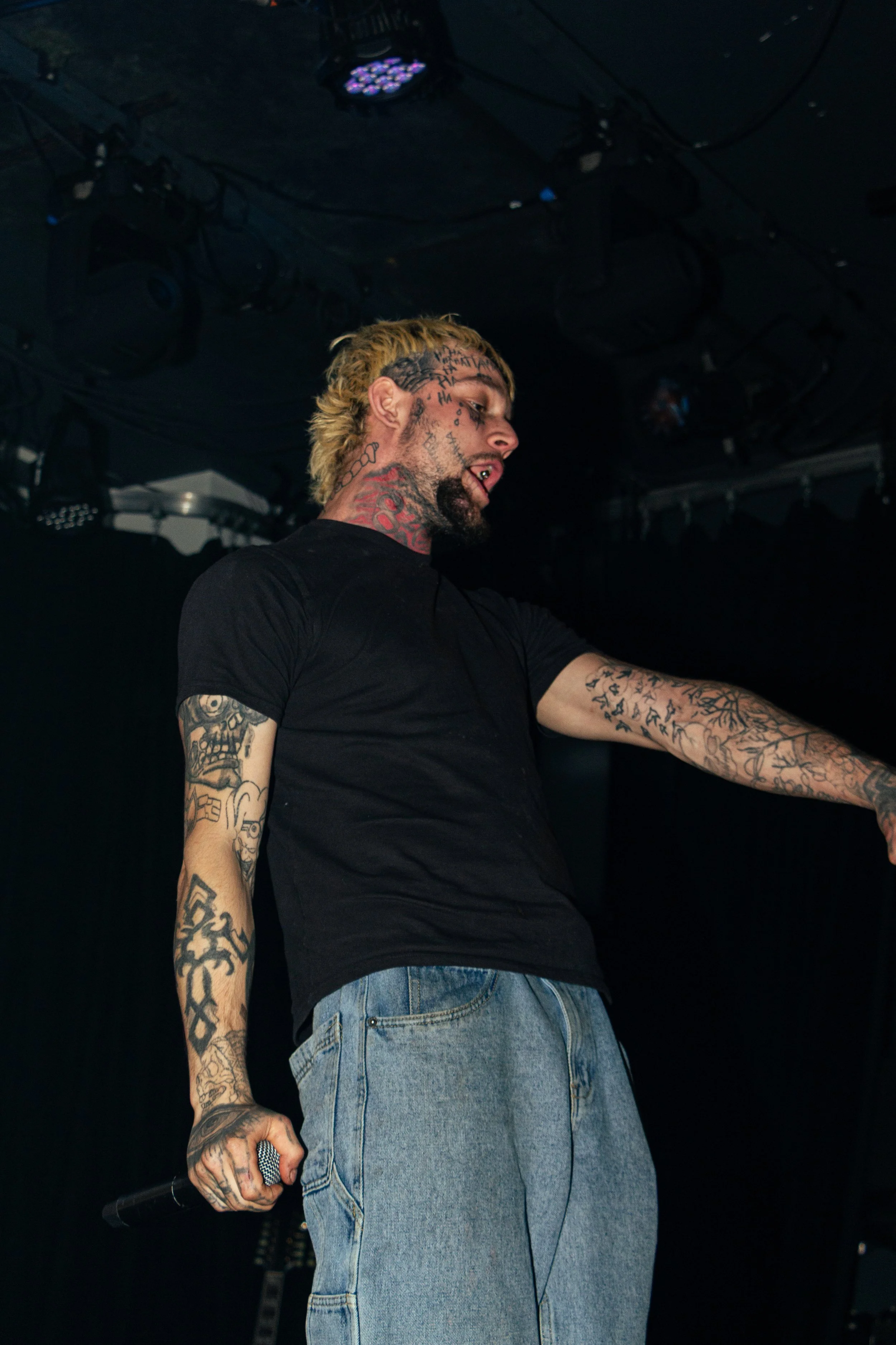 A tattooed man with blond hair performing on stage, wearing a black T-shirt and jeans, holding a microphone in his right hand, with stage lighting above him.