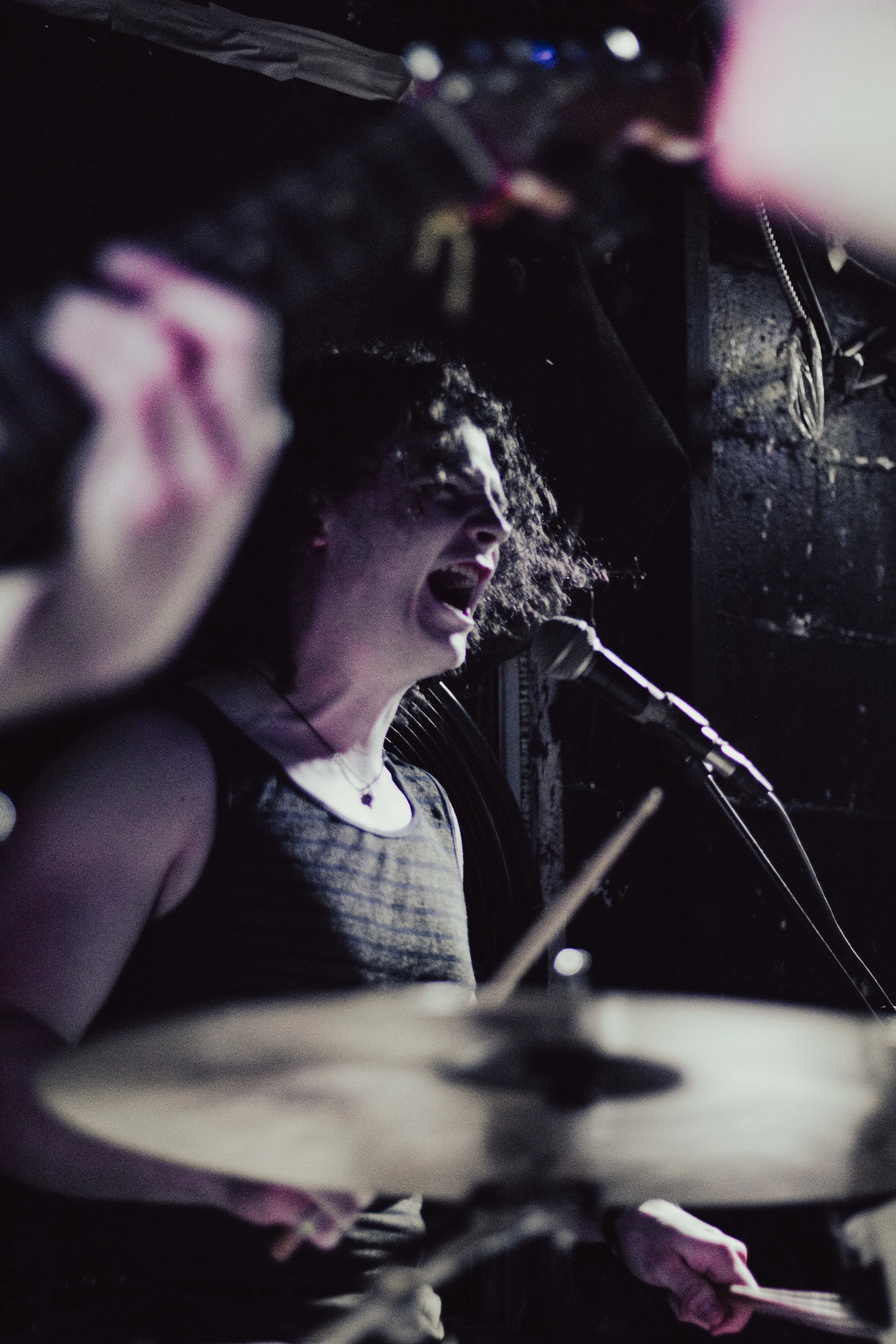 A young woman with curly hair shouting or singing passionately, with her mouth wide open, in a dark setting with equipment and dark background.