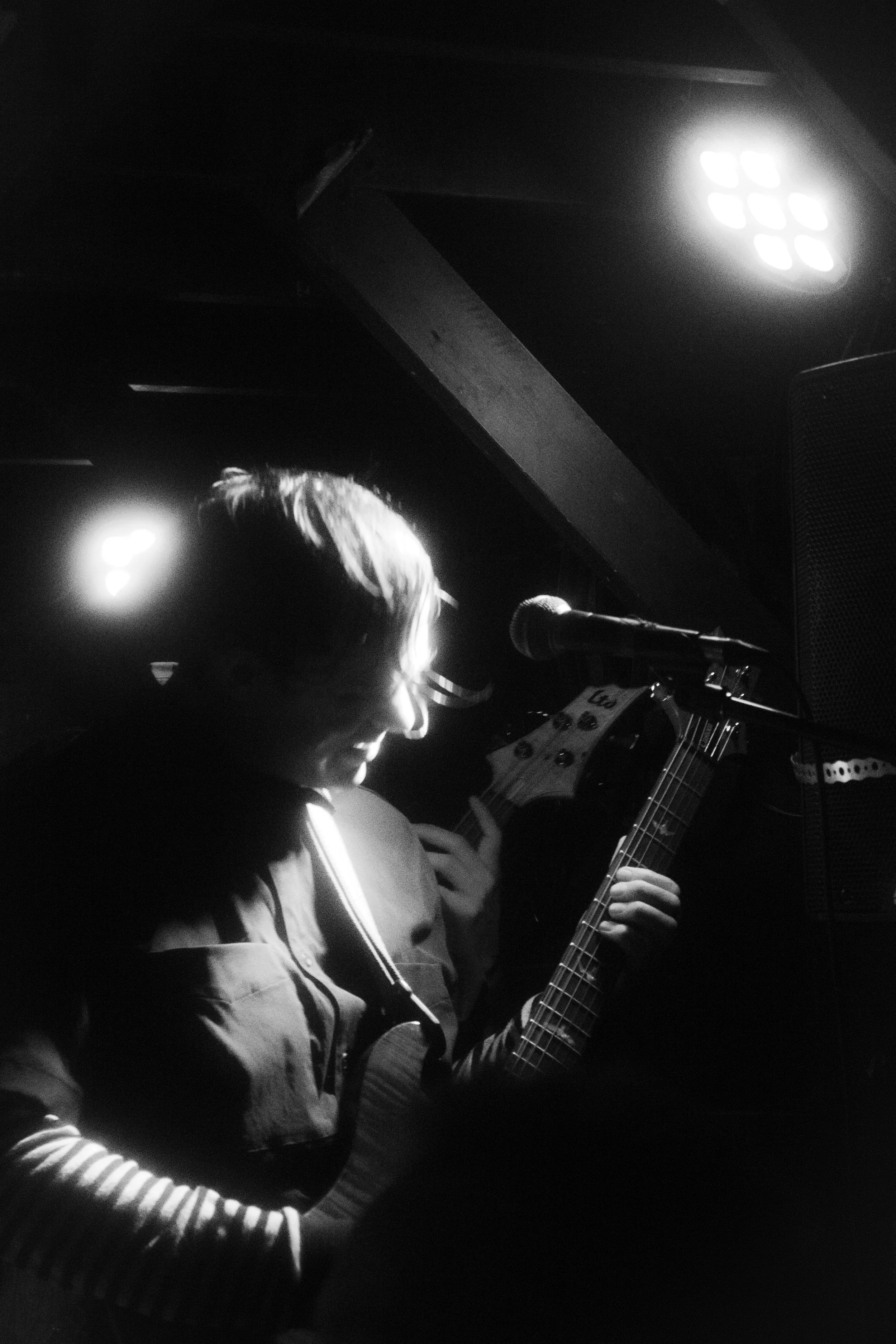 A black-and-white photo of a woman playing an acoustic guitar in a dimly lit space, with a microphone in front of her and bright lights overhead.