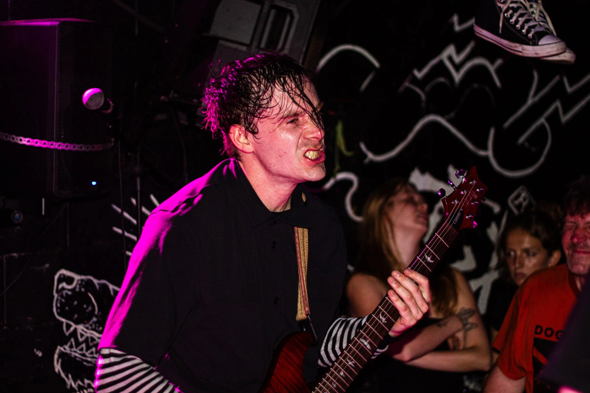 A male musician with wet hair and a black shirt playing an electric guitar at a live music venue, with a pink and purple stage light illuminating him. There are several audience members in the background.