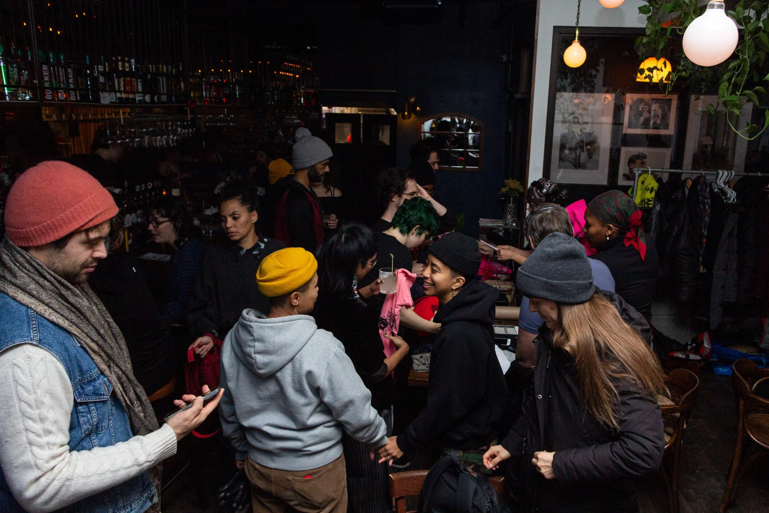 A group of people gathered in a dimly lit indoor space, possibly a bar or cafe, engaging in conversations. There are hanging lights and framed photographs on the walls, with various items like jackets and bags around.
