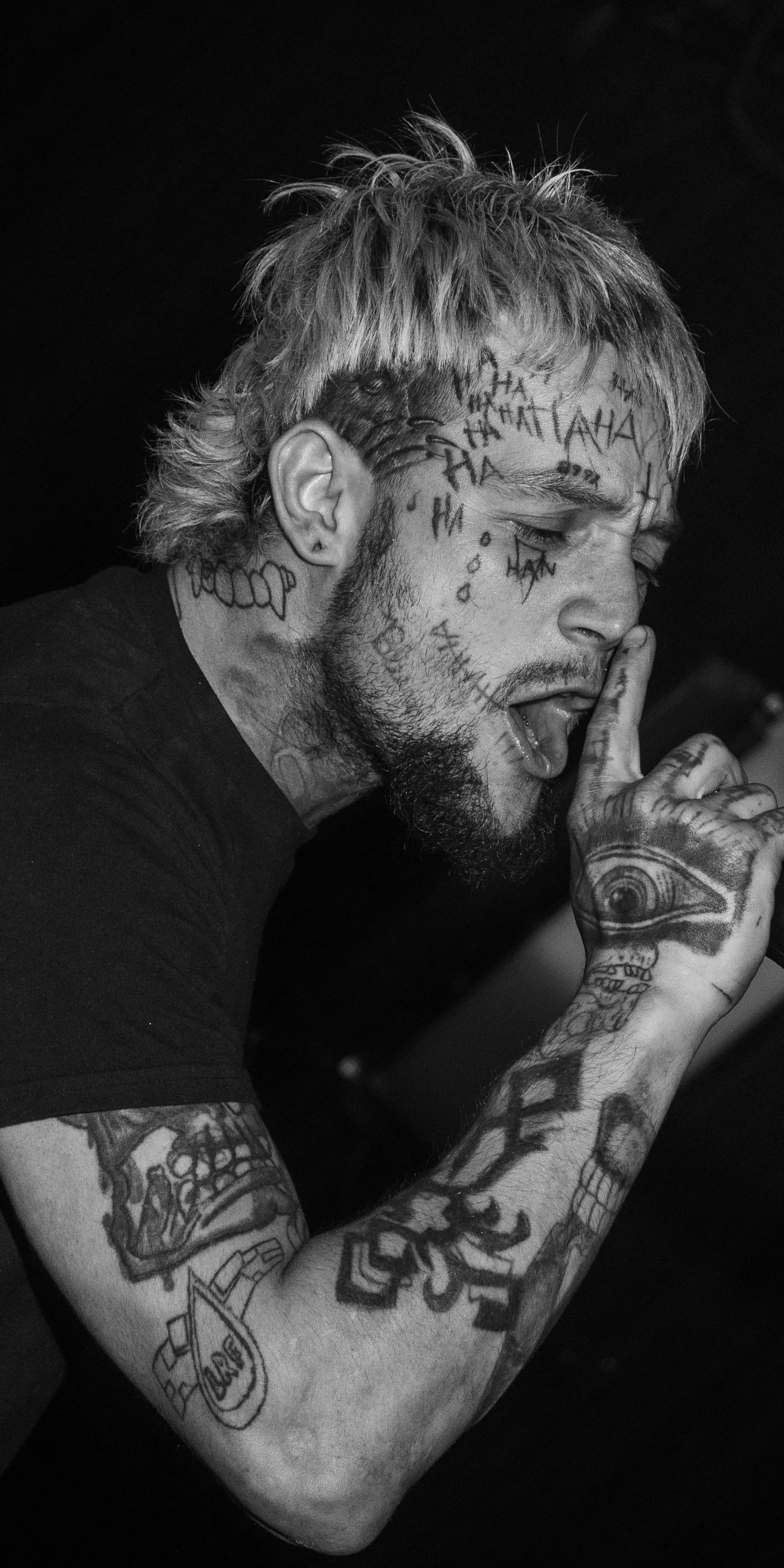 Black and white photo of a tattooed man with light-colored hair, tattoos on his face, arm, and hand, with eyes closed.