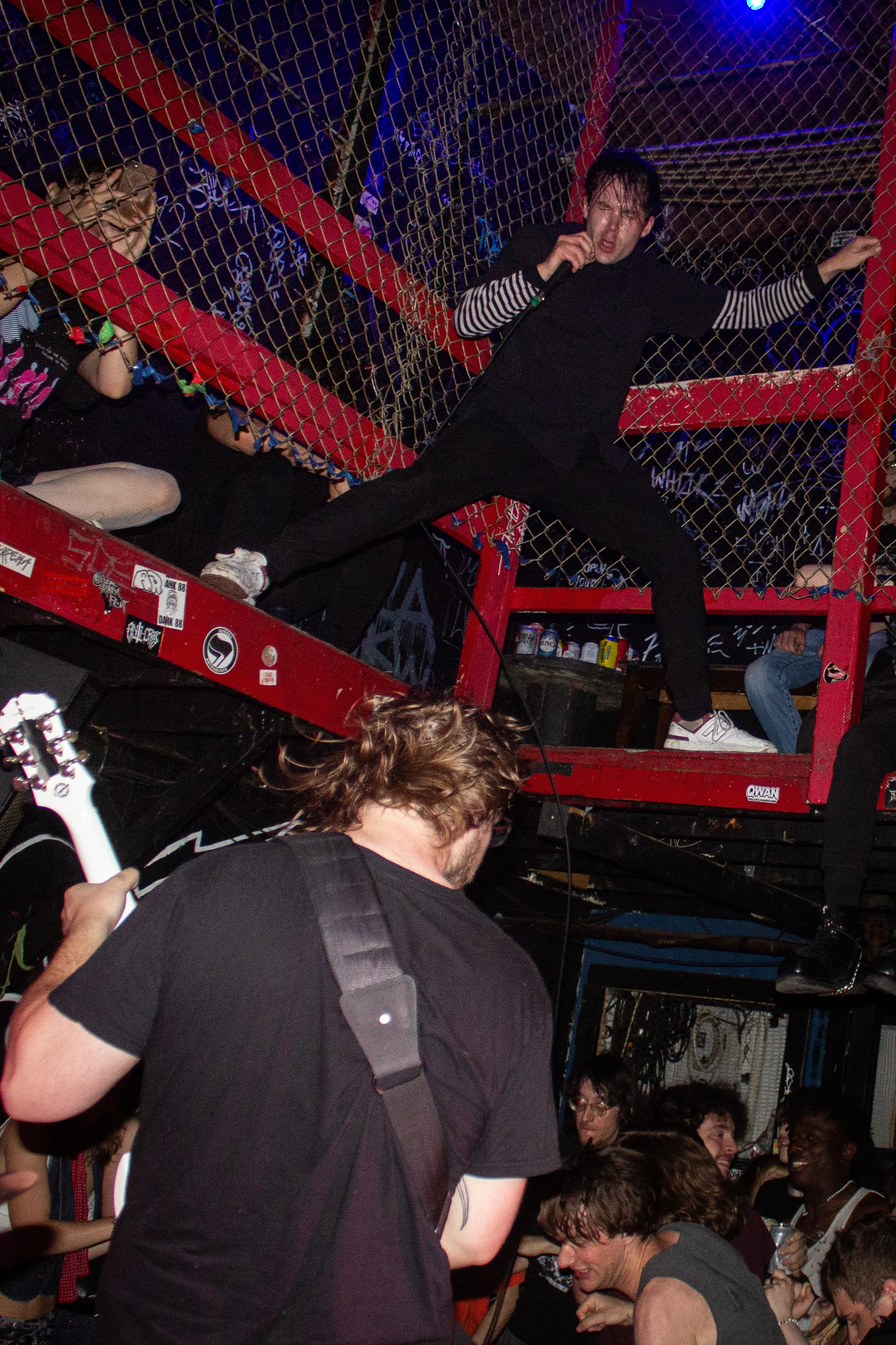 A man in a black shirt and striped long-sleeve shirt holding a microphone, with his left arm extended and a wide stance in front of a crowd inside a venue with graffiti and neon signs. Multiple other people are seen smiling and talking, some with gla
