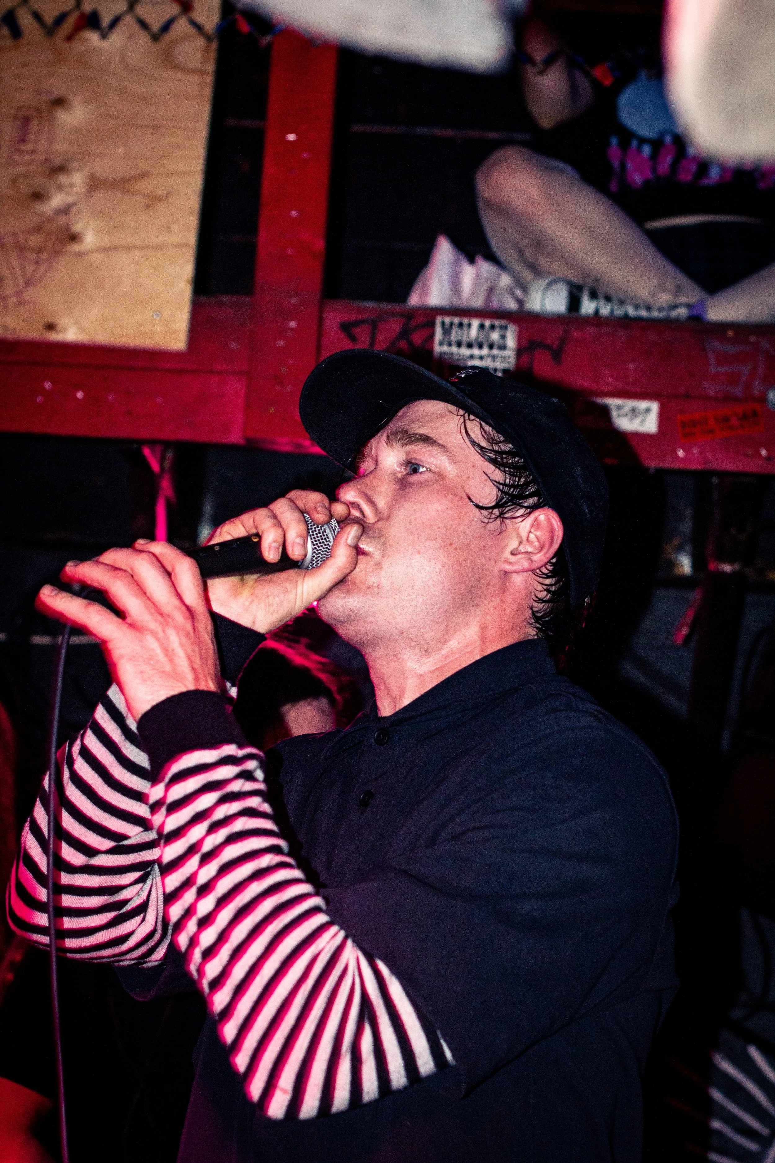 A man wearing a black hat and black shirt singing into a microphone, partially obscured by a person in a striped shirt. The background includes a red-painted surface with stickers and graffiti.