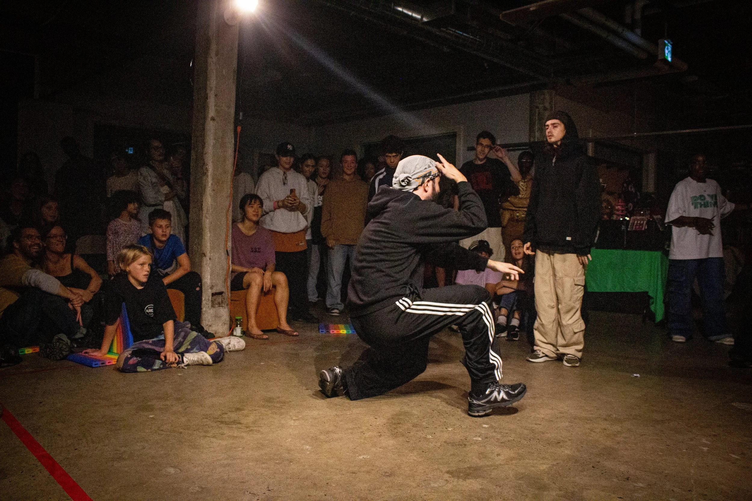 A group of people watching a performer kneeling down and gesturing during a performance in a dimly lit indoor space.