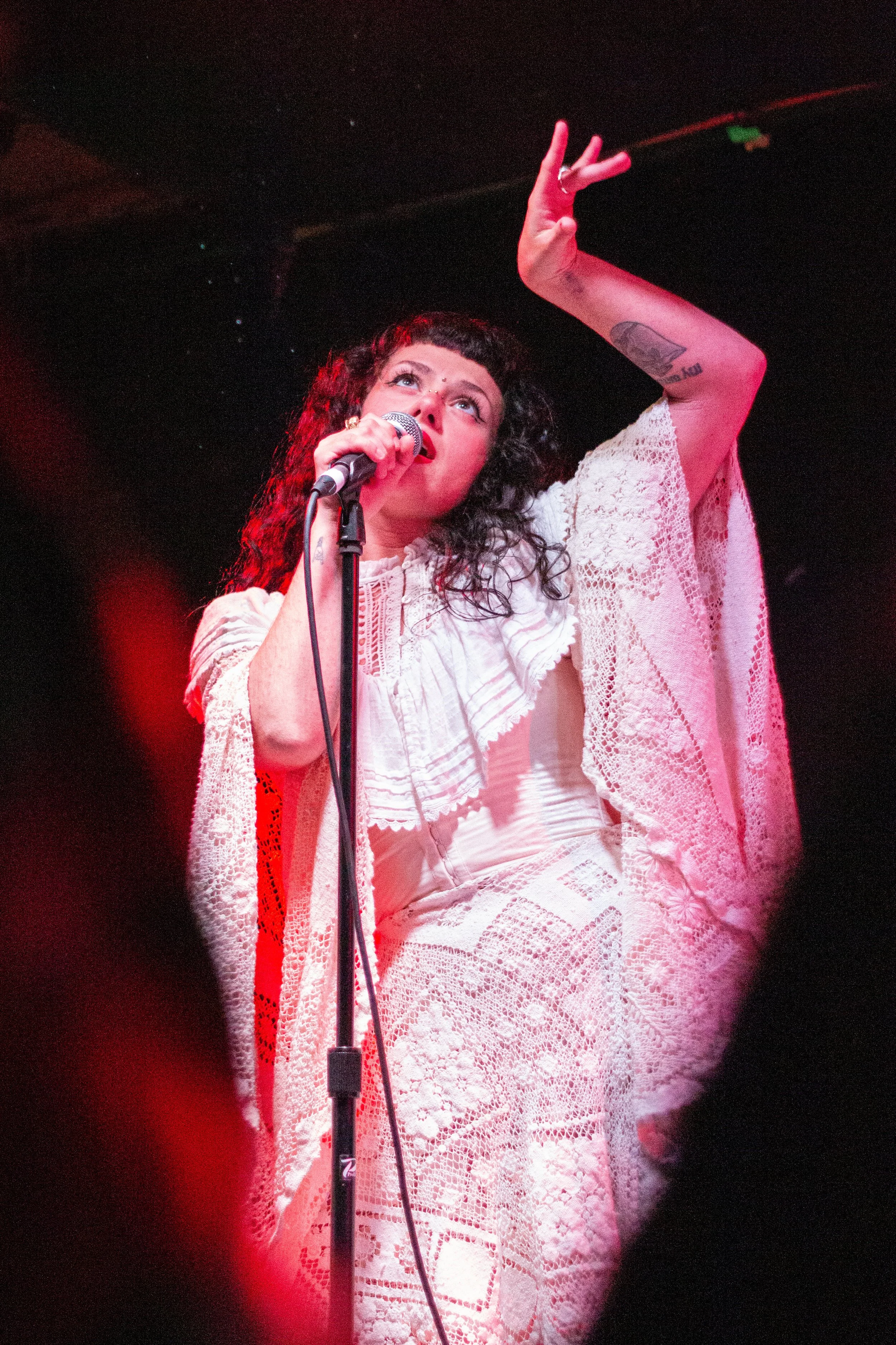 A woman with curly dark hair and tattoos on her arm is singing or speaking into a microphone on stage, raising her hand with fingers in a peace sign, wearing a white lace dress.