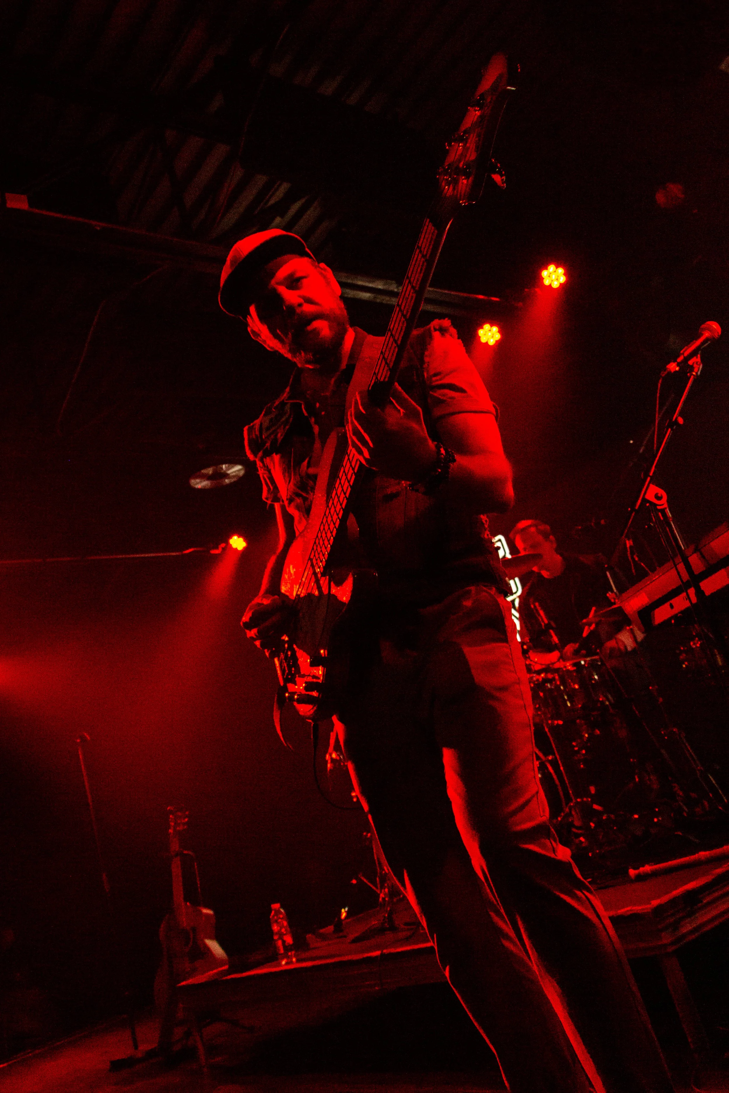 A male musician wearing a red bandana and a vest playing an electric guitar on stage, illuminated by red stage lighting. A keyboard player is visible in the background, and microphones are set up nearby.